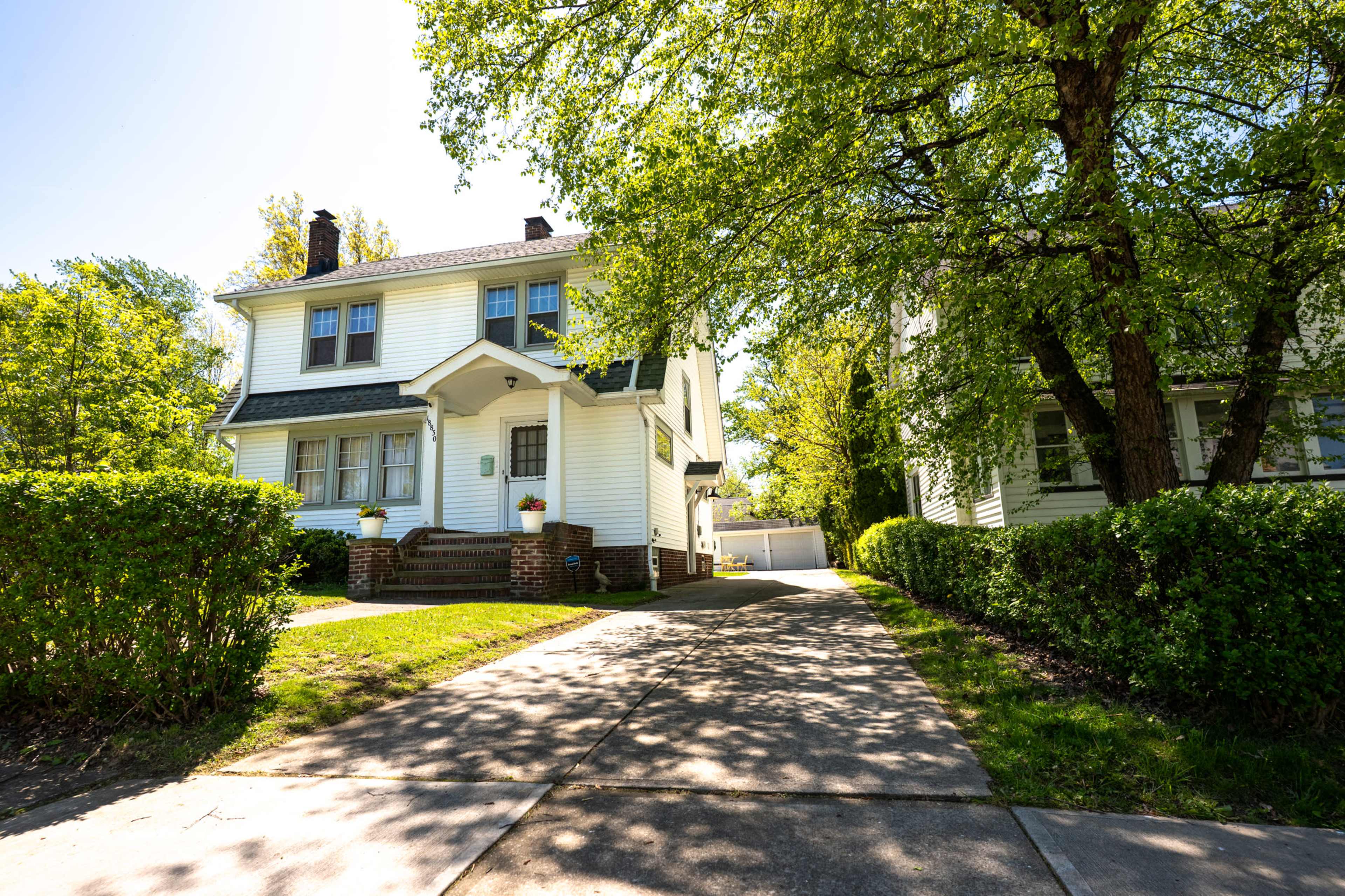A two-story white house with a front porch is surrounded by well-maintained greenery and a concrete driveway.