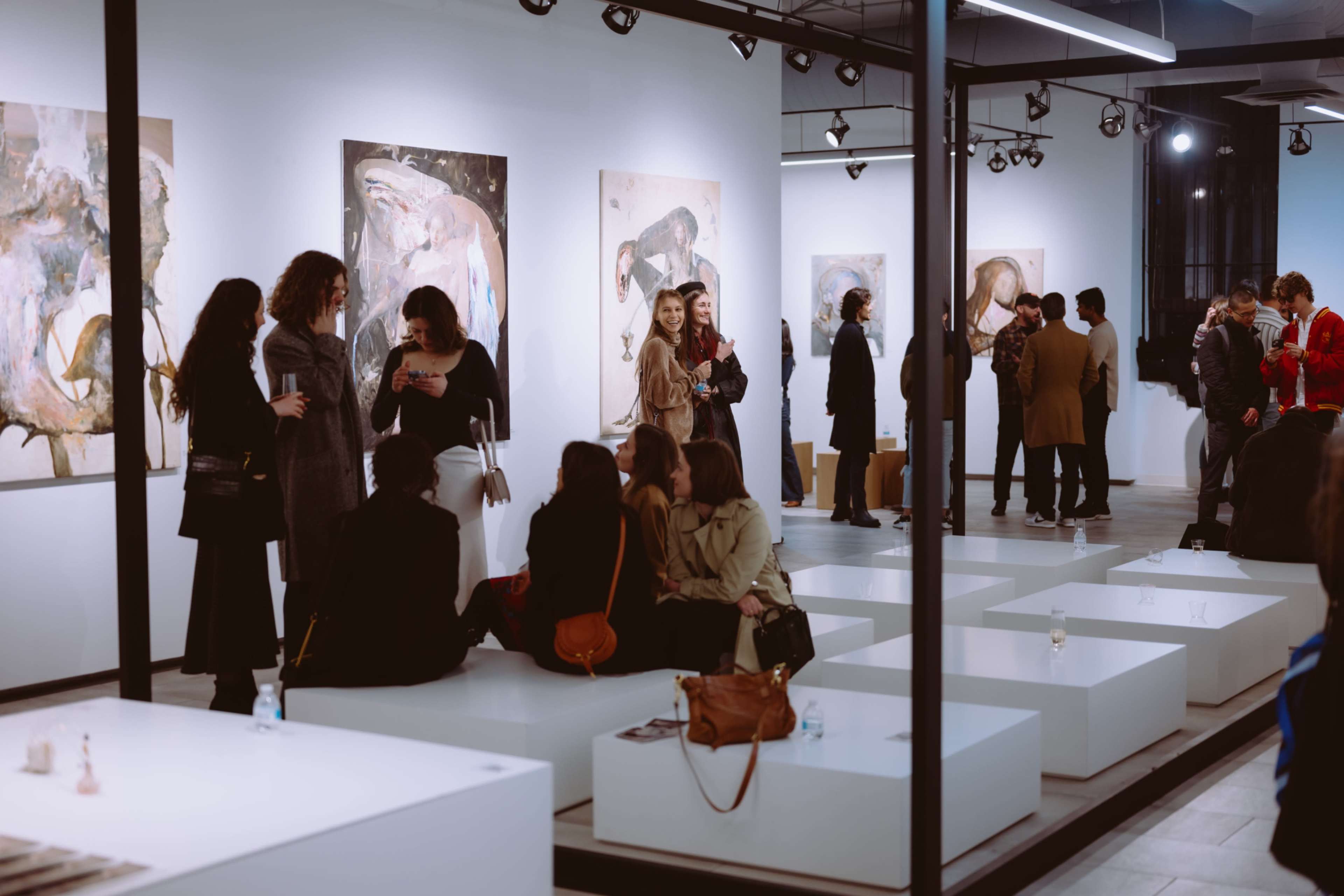 People gather and socialize in an art gallery showcasing large paintings on the walls.