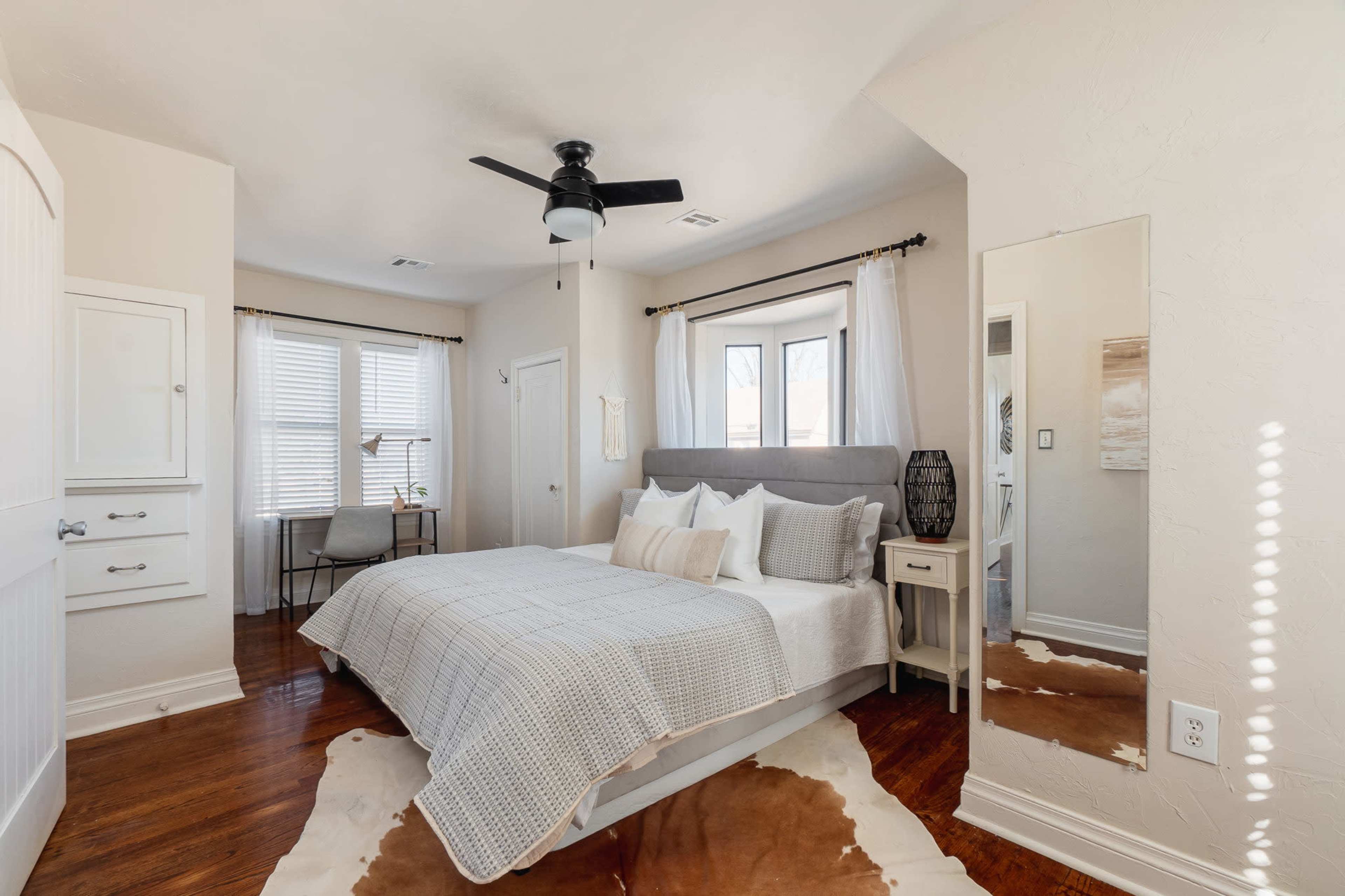 A neatly arranged bedroom featuring a bed, a small nightstand, a fan, and a desk beside large windows.