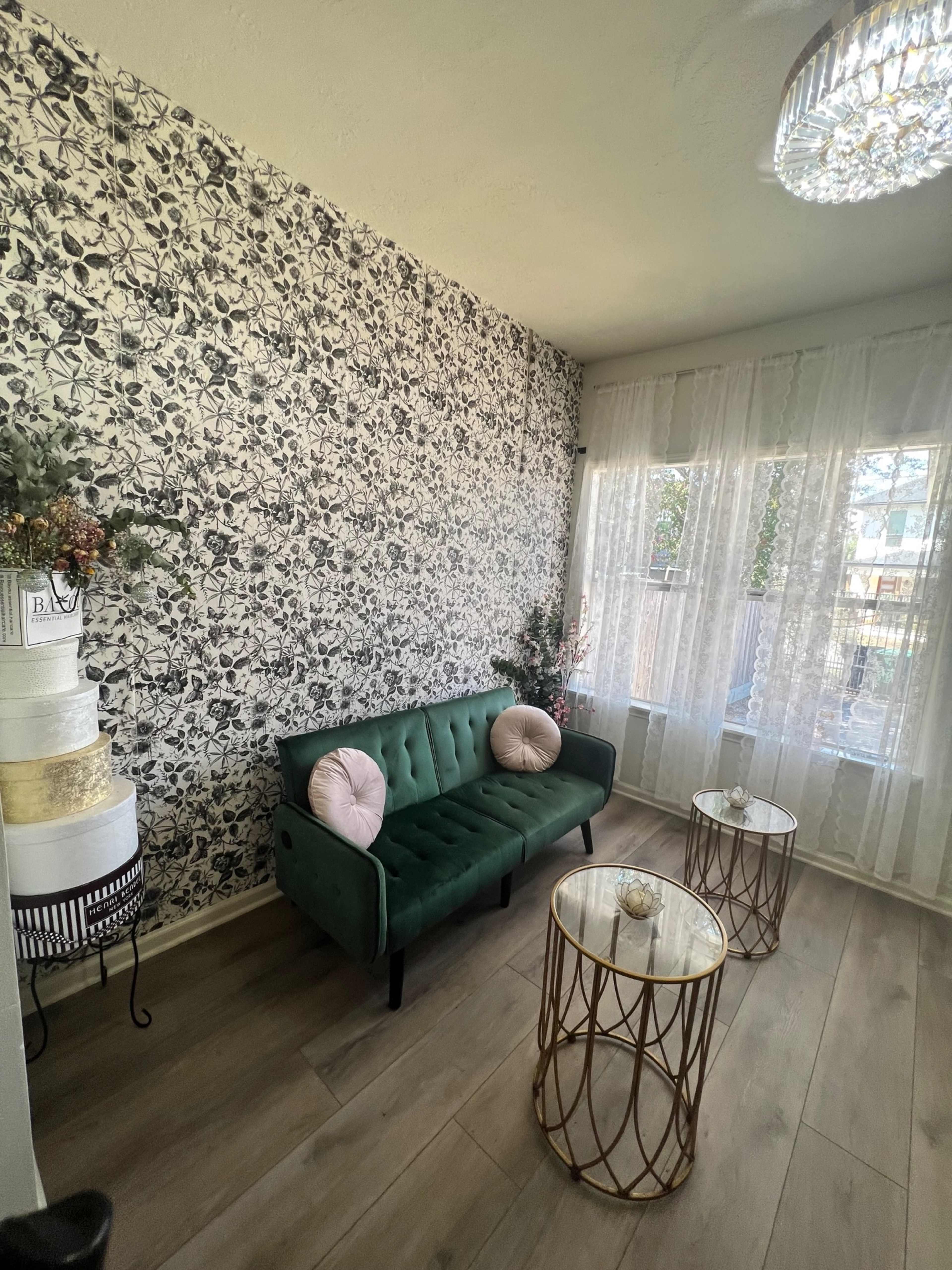 The image shows a stylish room with a green sofa, two round gold coffee tables, and a floral patterned wall, illuminated by a chandelier.