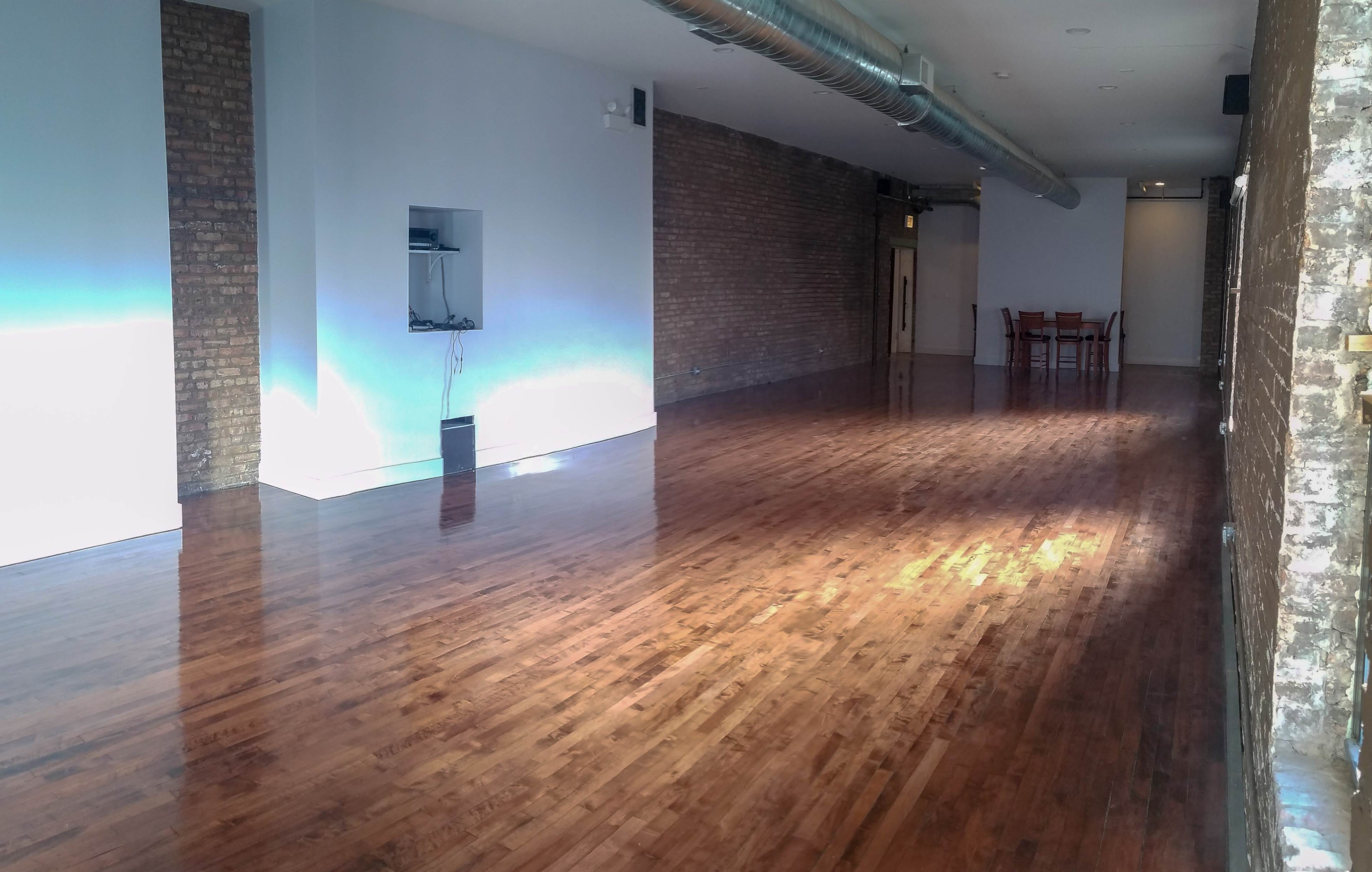 The image shows a spacious, empty room with polished wooden floors, exposed brick walls, and a dining table set in the background.