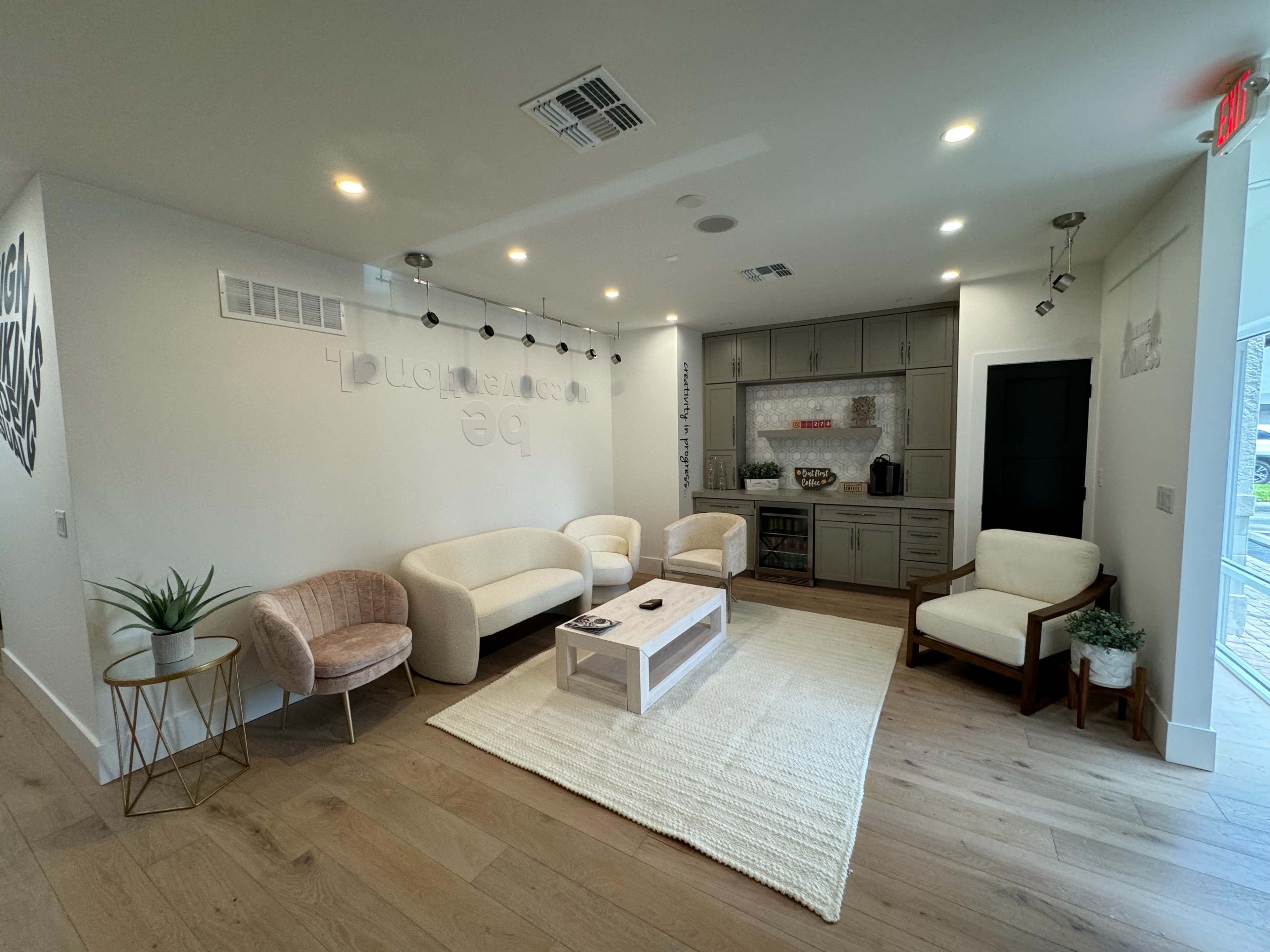 A modern waiting area featuring a light-colored sofa set, a coffee table, and a rug on a wooden floor, with a kitchenette visible in the background.