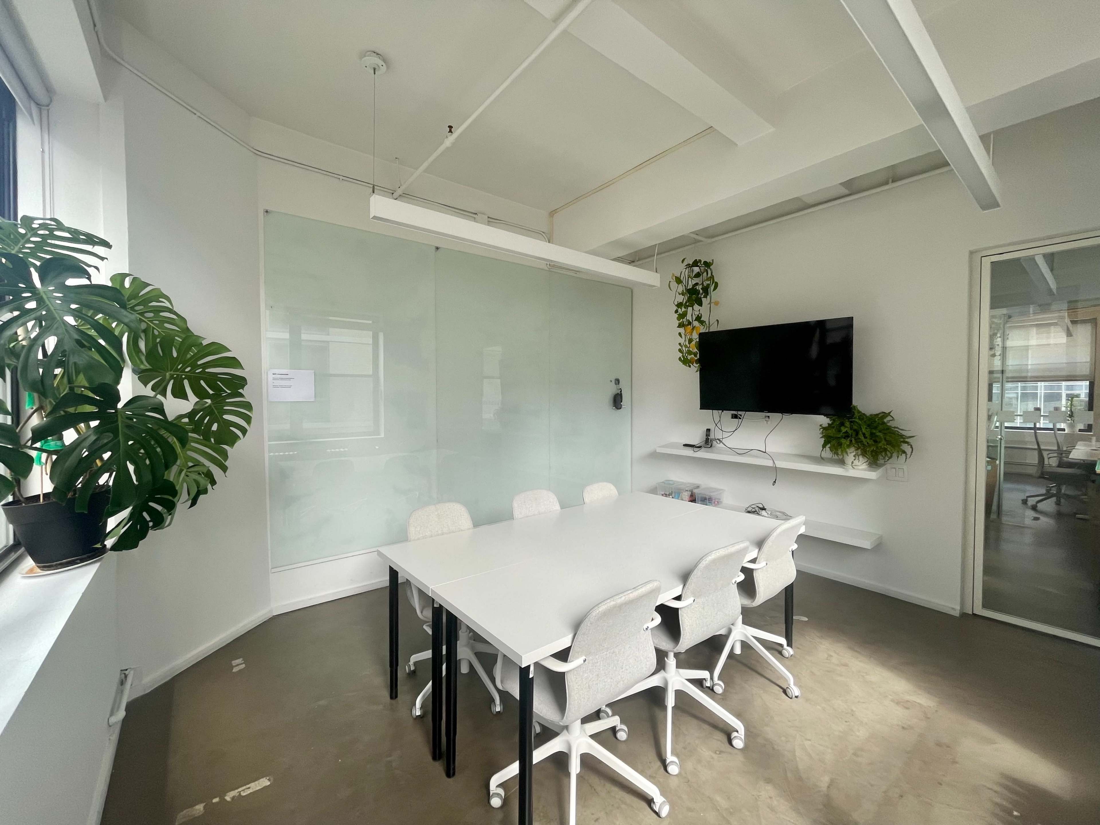 A modern conference room with a large white table surrounded by ergonomic chairs, a wall-mounted TV, and potted plants near the windows.