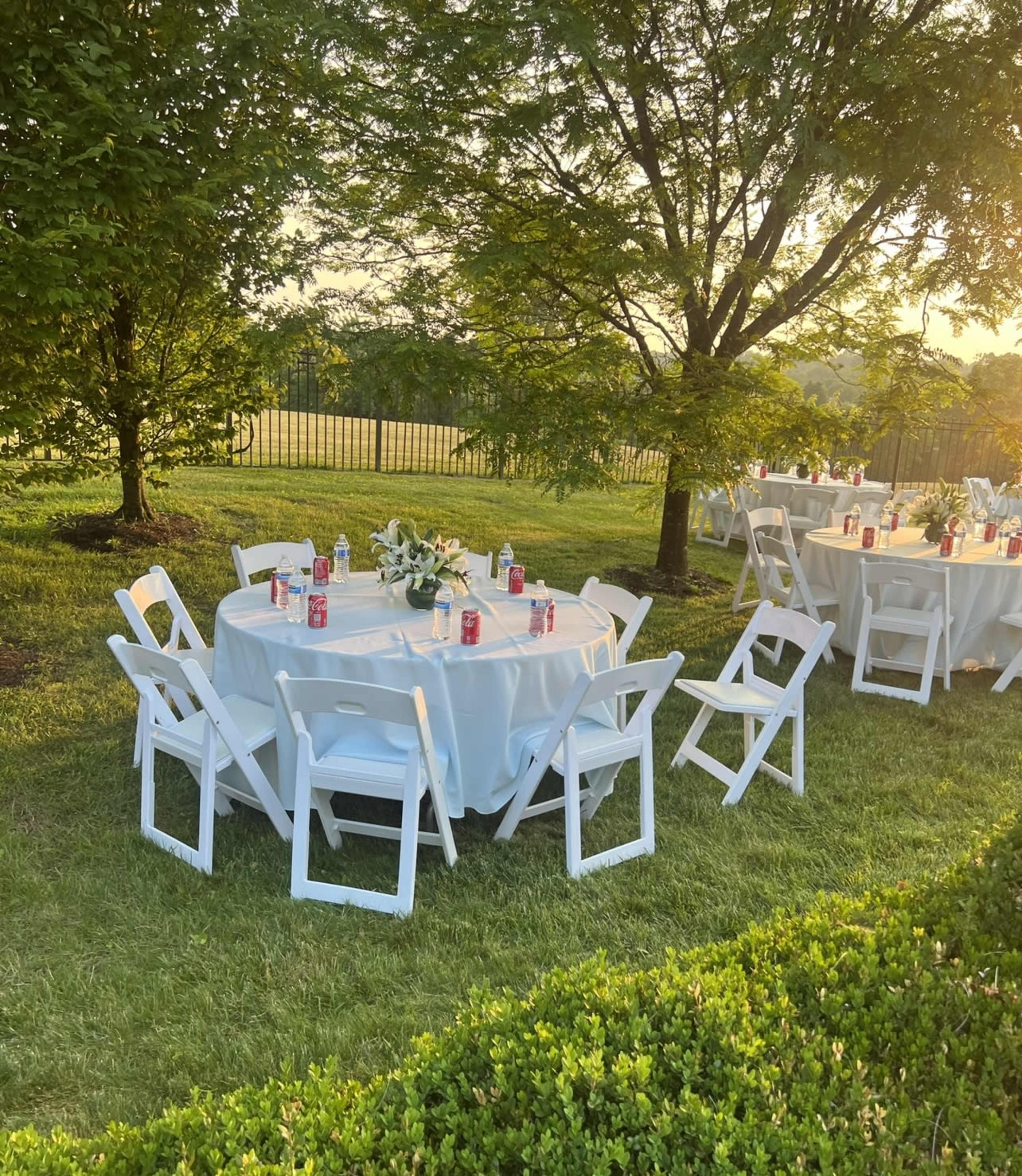A set of round tables with white tablecloths and chairs is arranged in a grassy area, surrounded by trees and a fence in the background.