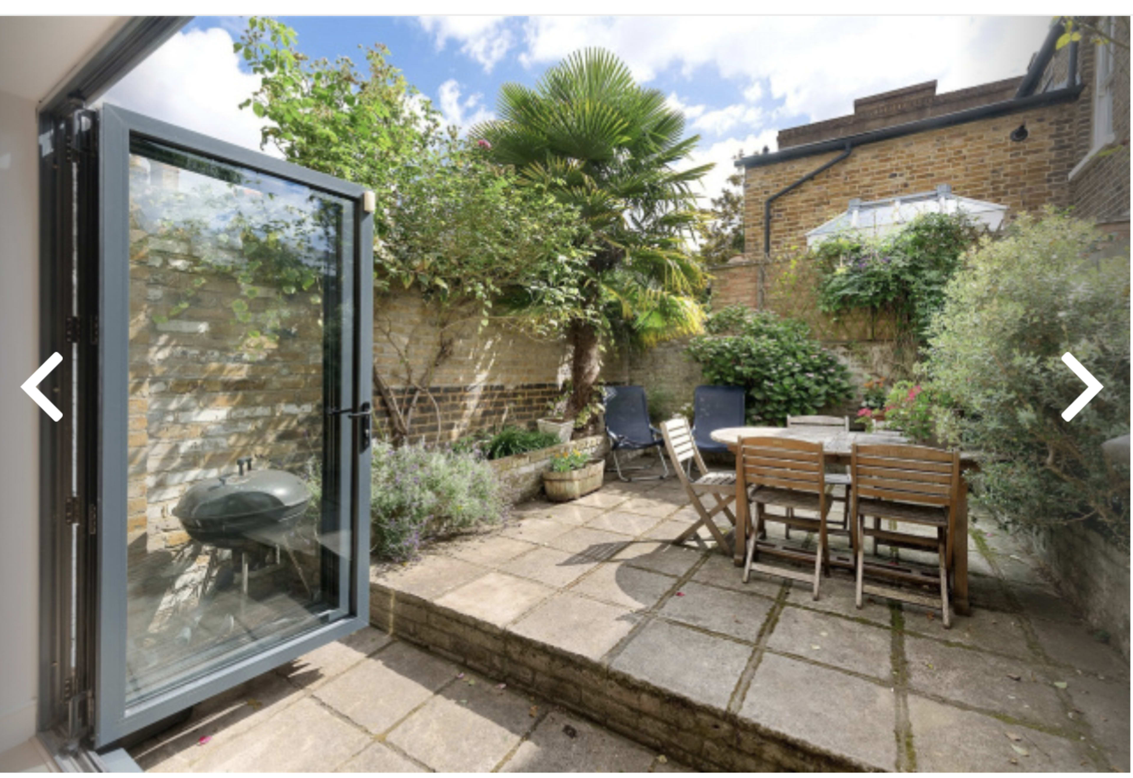 The image shows a small outdoor patio area with a table and chairs, surrounded by greenery and a grill, accessed through a glass door.