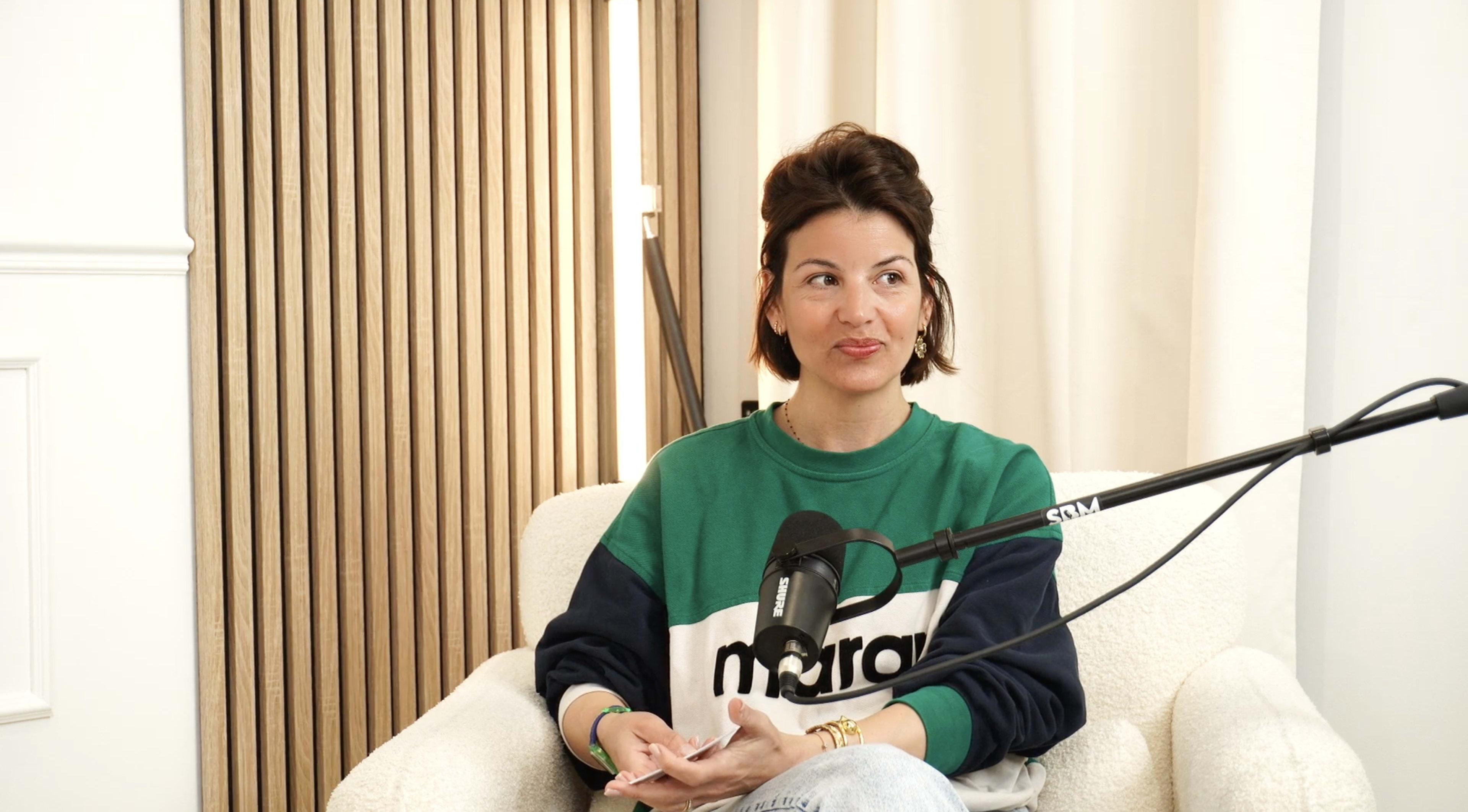 A person sits on a white chair, wearing a green and navy sweater, with a microphone positioned nearby in a well-lit room featuring wooden paneling.