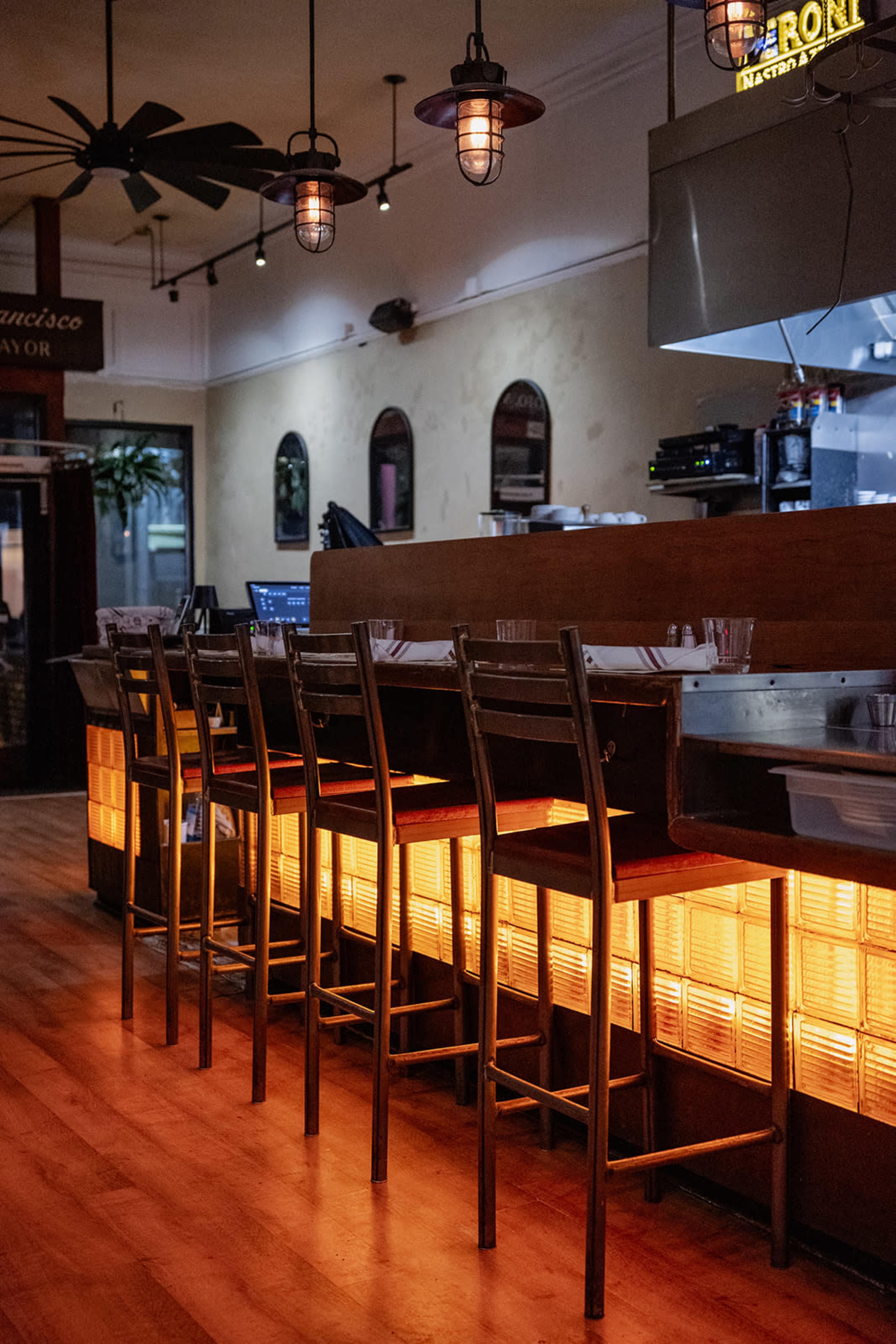 The image shows a row of metal chairs lined up at a bar with warm, glowing lights underneath and a kitchen area in the background.