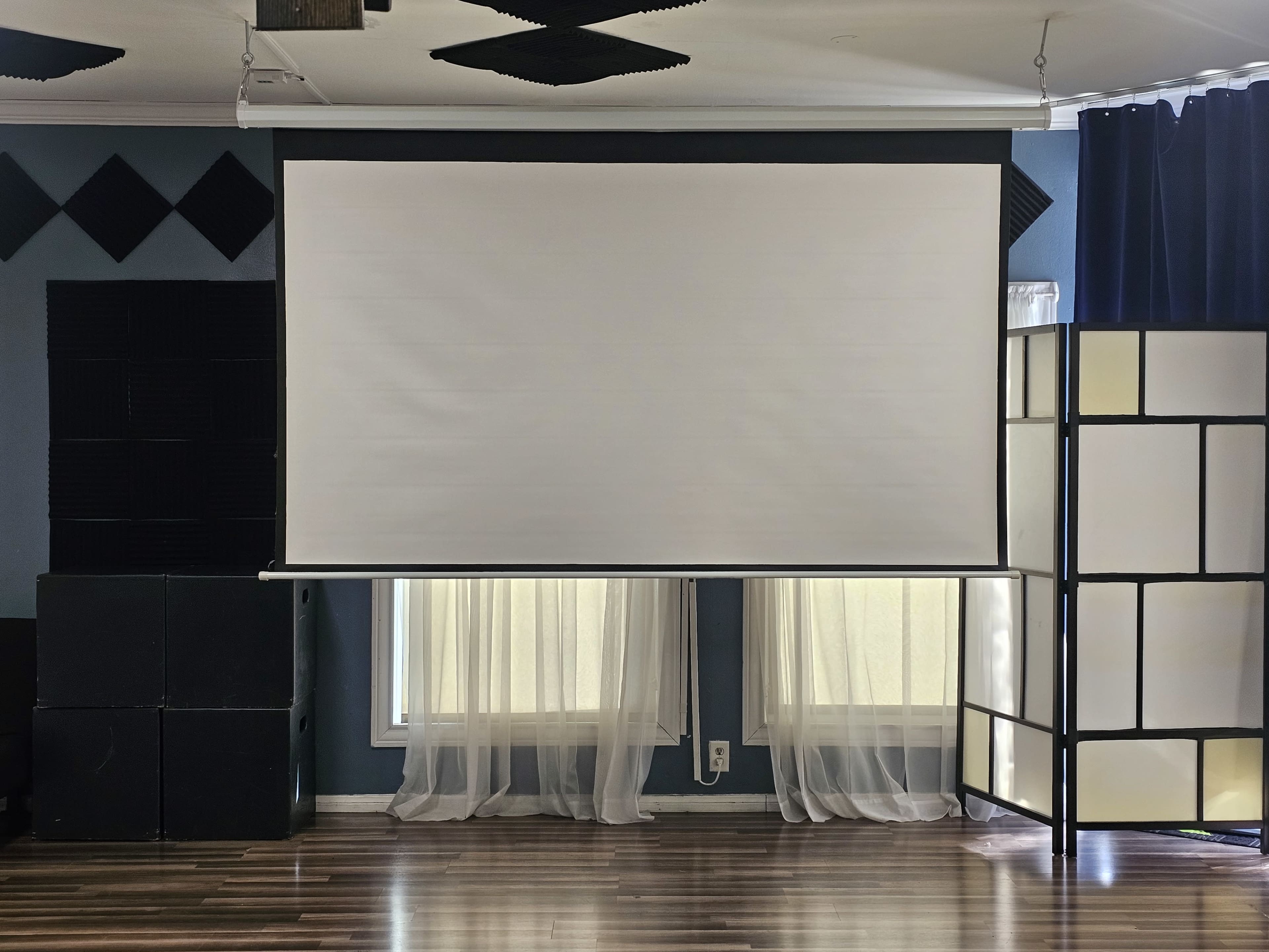 The image shows a blank projection screen hanging in a room with wooden flooring, soft curtains, and acoustic panels on the walls.