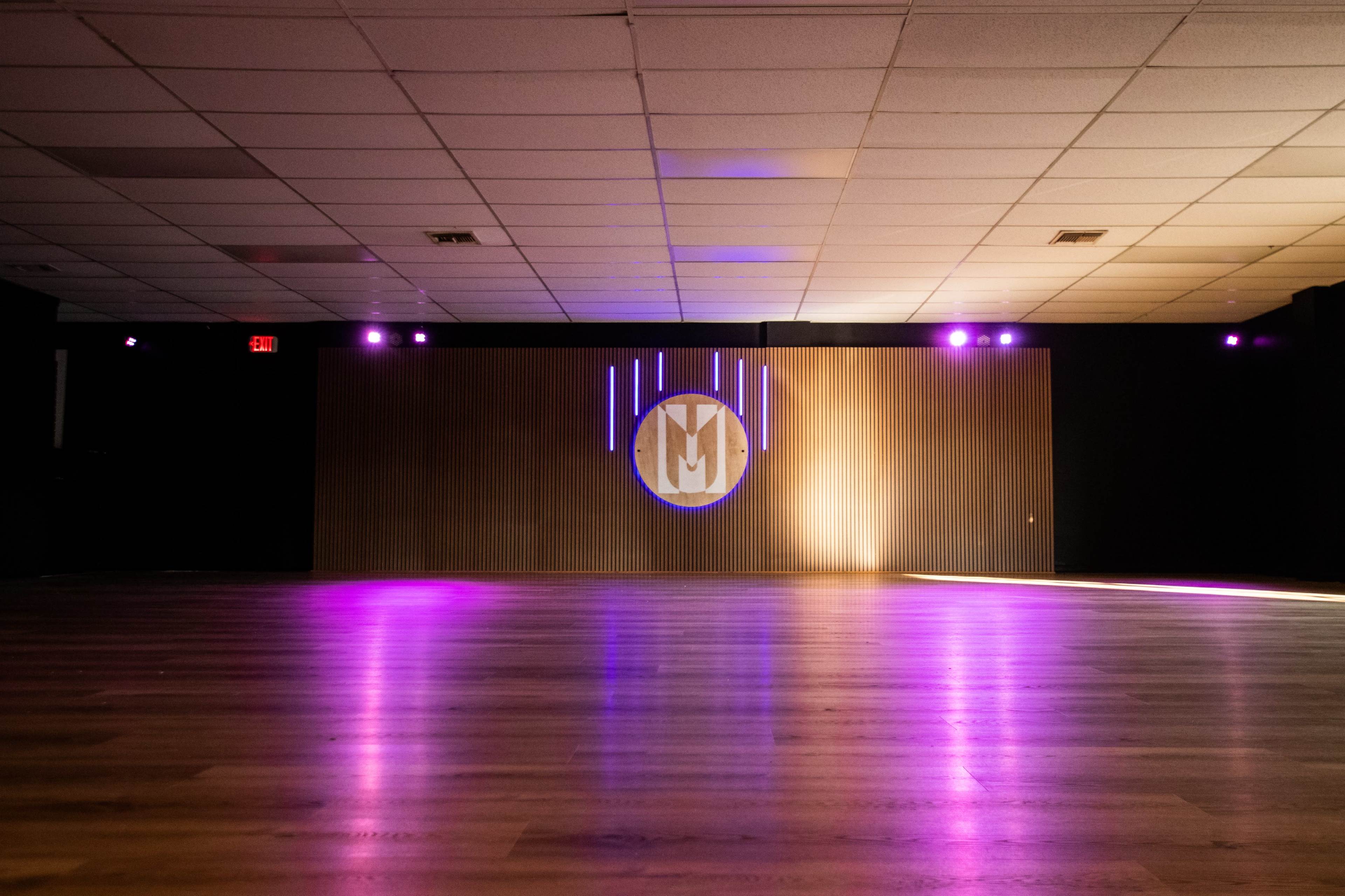The image shows an empty event space with a wooden wall featuring a lighted logo, illuminated by purple and white lights.