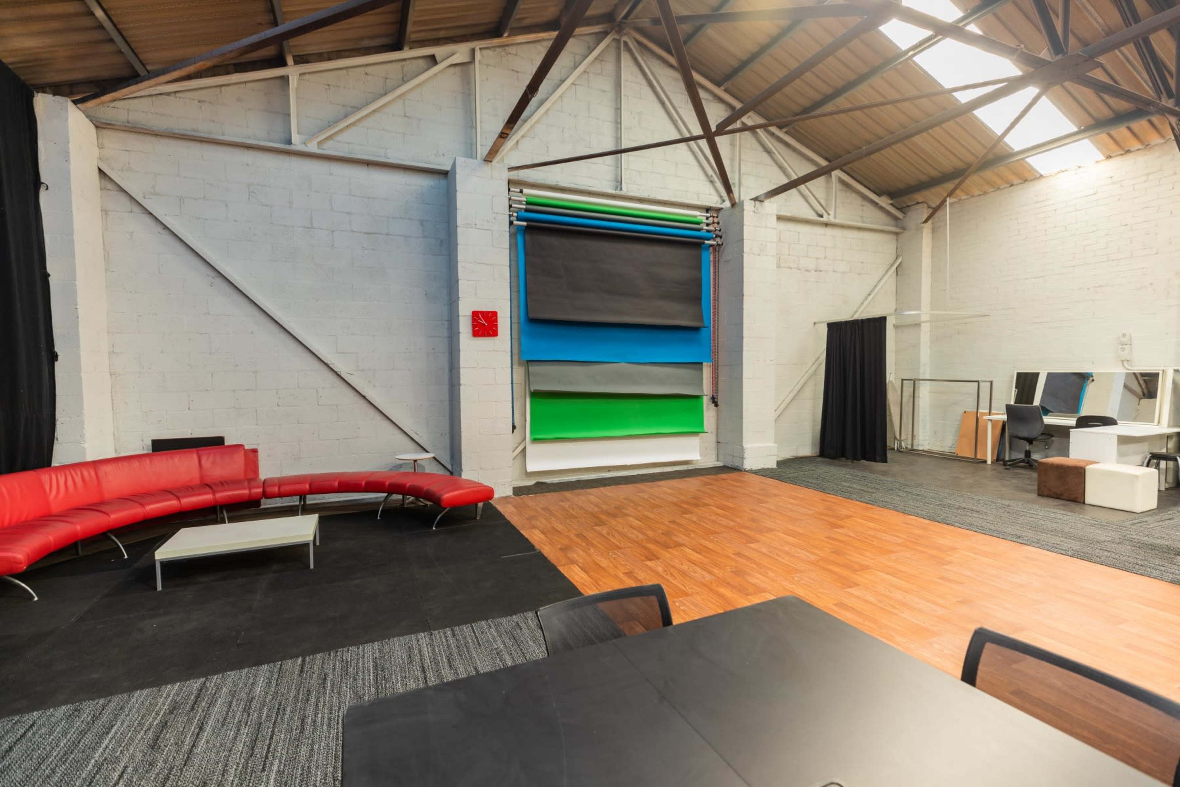 A spacious indoor area with a red curved sofa, light wood flooring, and a backdrop support featuring several solid color rolls in blue, black, green, and gray.