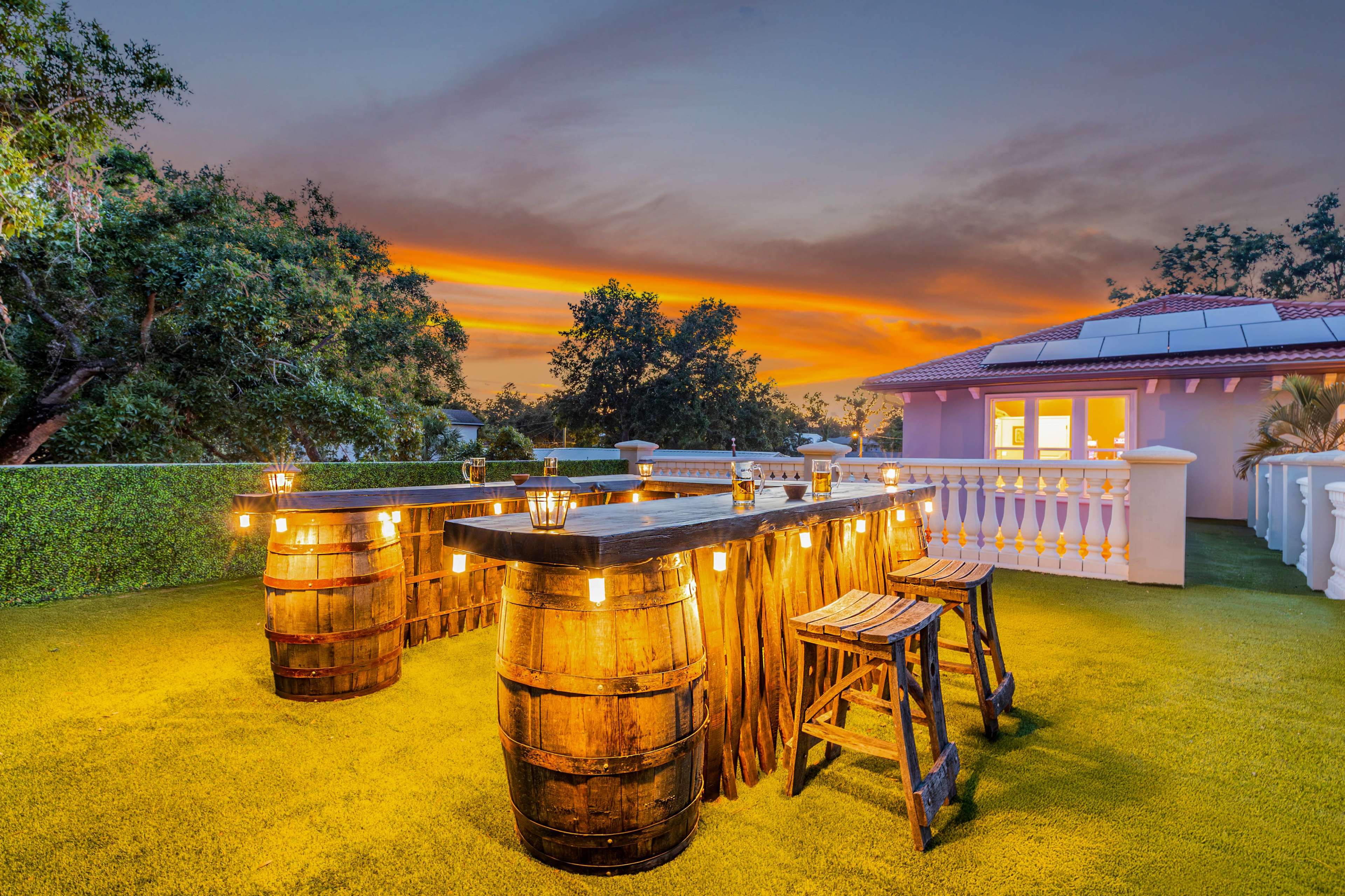 The image shows a rooftop bar made of wooden barrels, illuminated by string lights, with a sunset backdrop featuring colorful clouds.