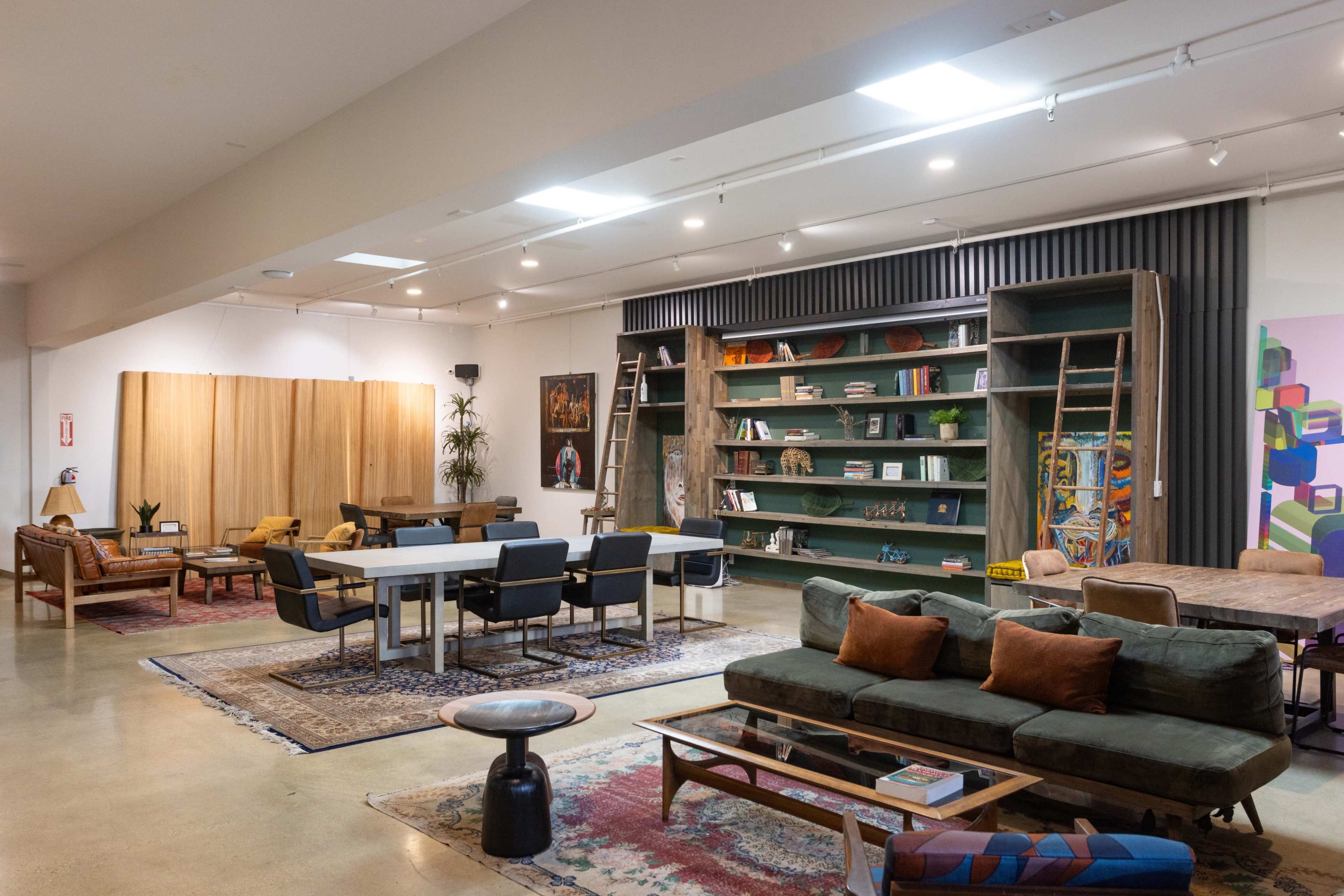 The image shows a spacious interior featuring various seating areas, a large bookshelf with ladders, and a dining table, all set against a backdrop of artwork and plants.