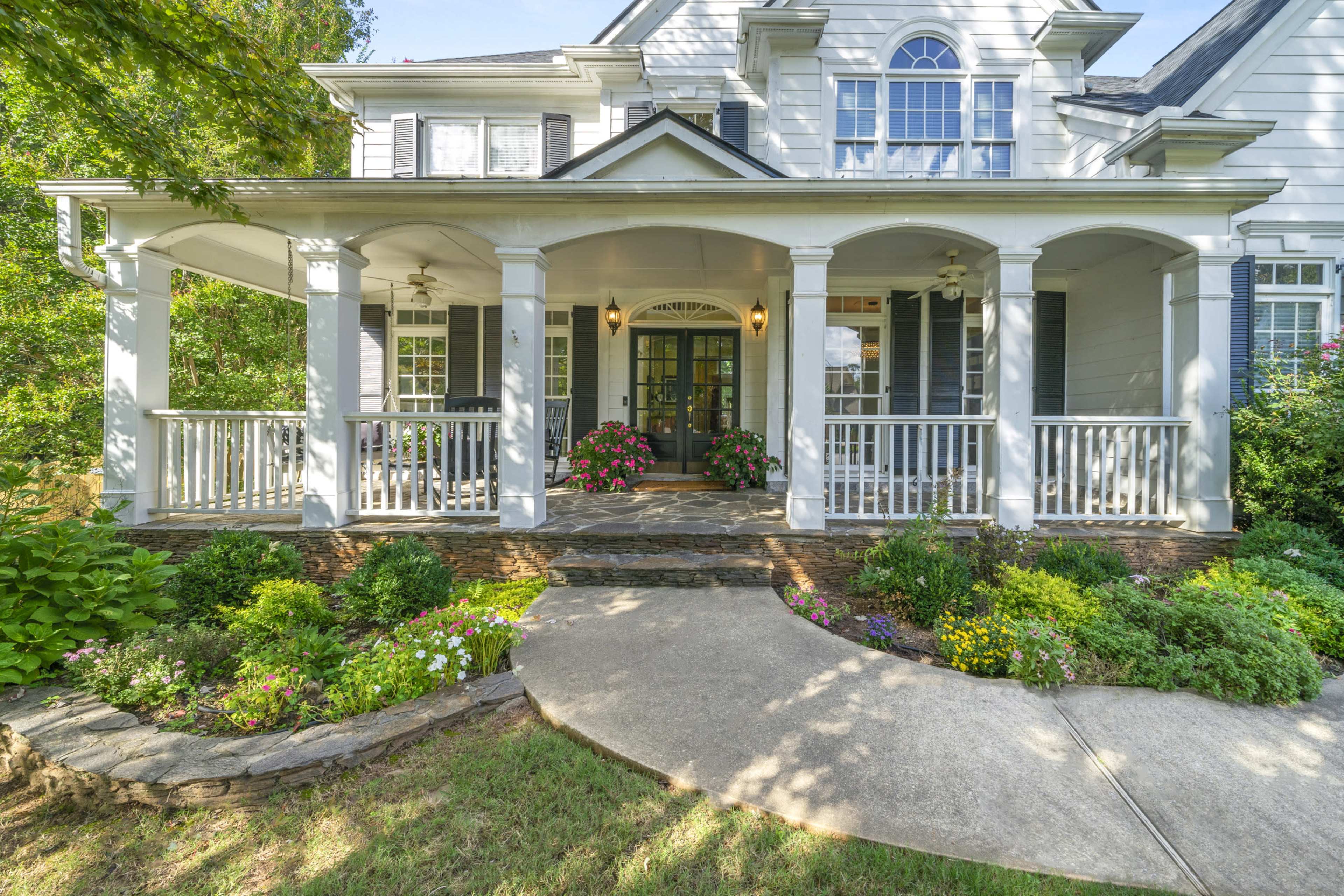 The image shows a two-story house with a covered front porch, stone walkway, and landscaped flower beds.