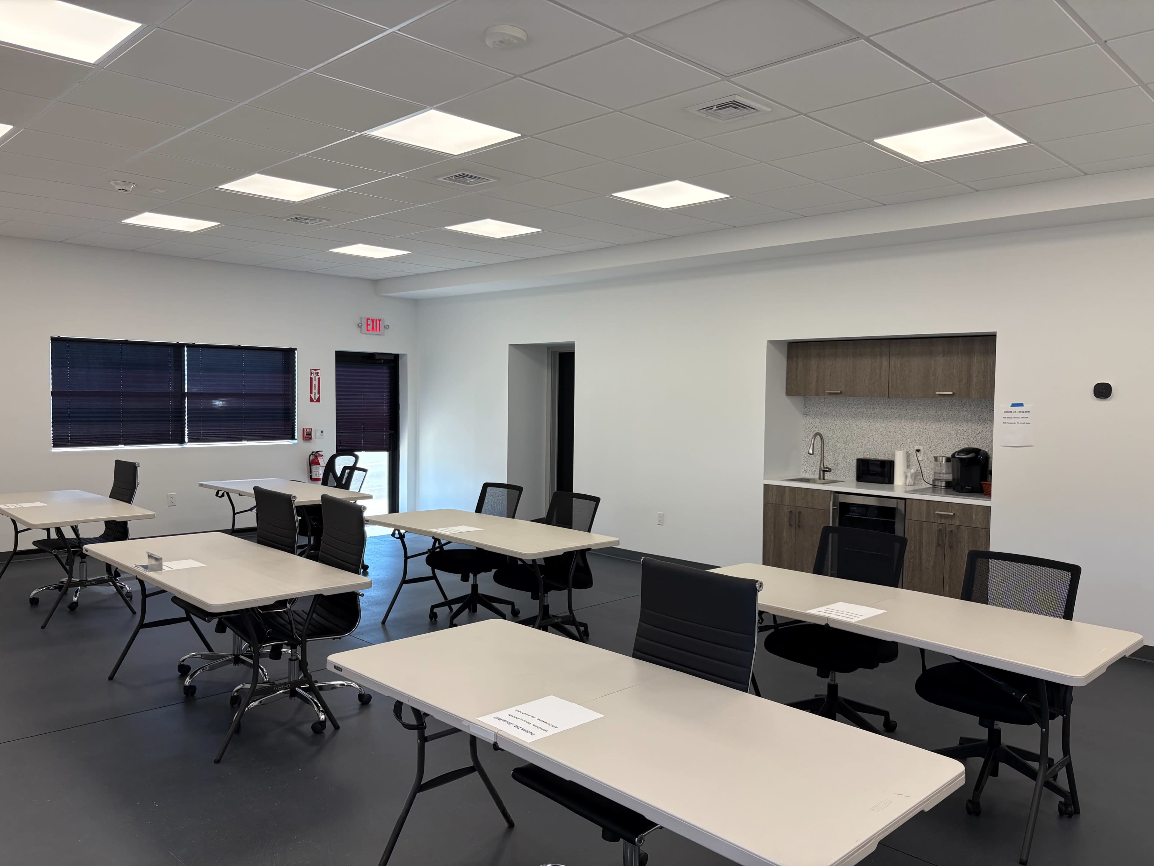 A spacious meeting room is set up with several tables and chairs, featuring a kitchenette in the corner.