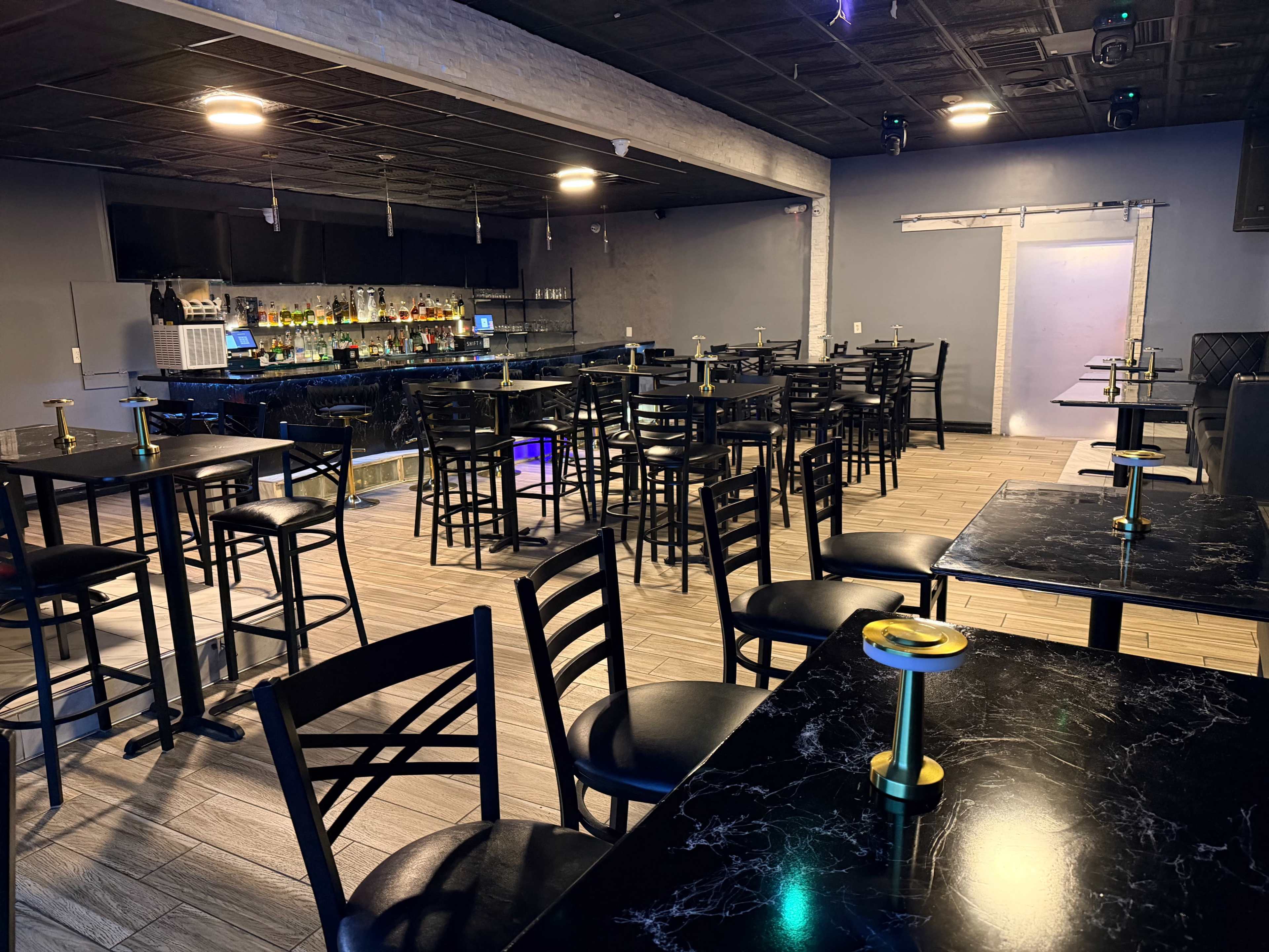 The image shows a nearly empty bar area with black tables and chairs arranged around a dimly lit space featuring a bar in the background.
