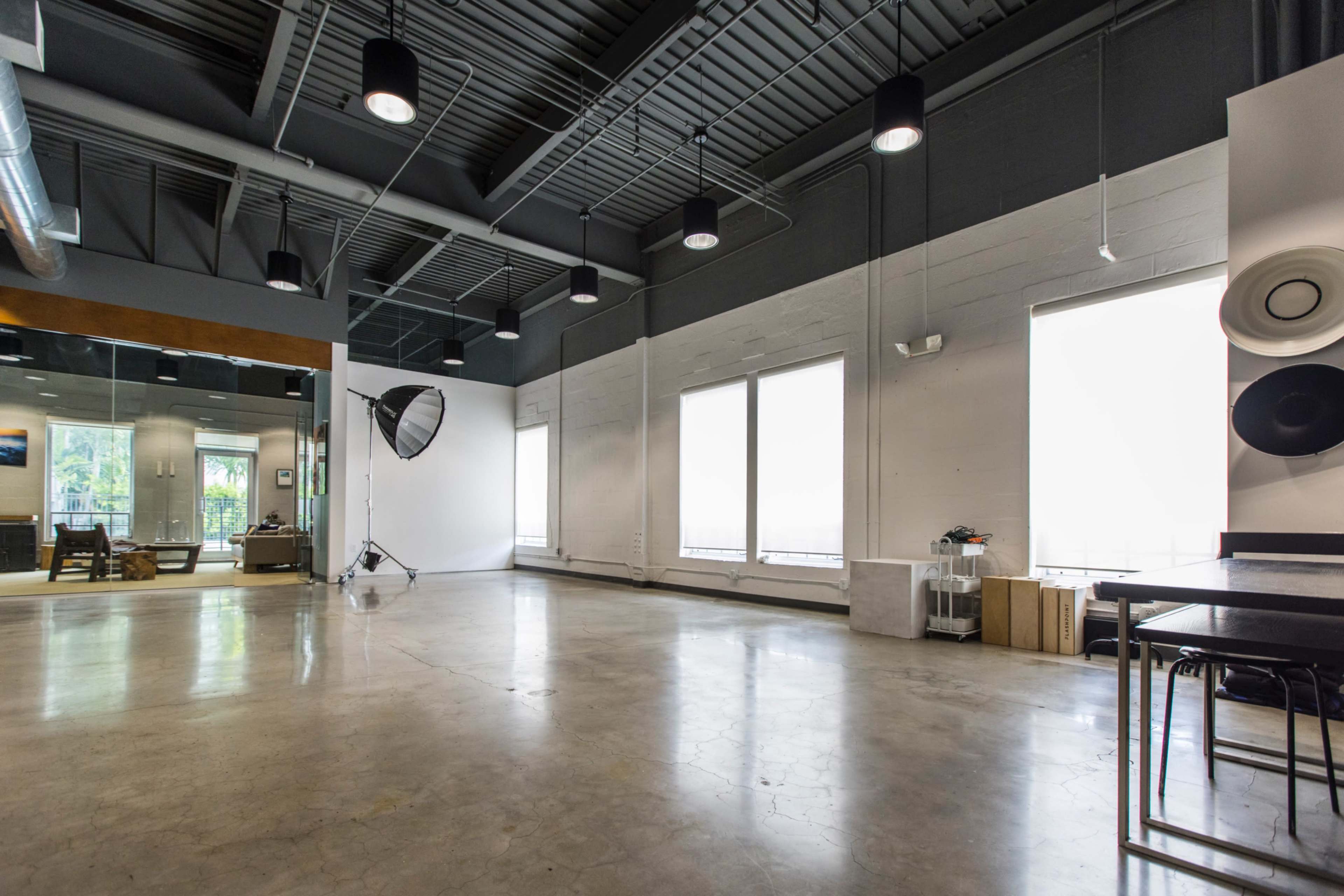The image shows a spacious, modern studio with large windows, concrete floors, and overhead lighting equipment.