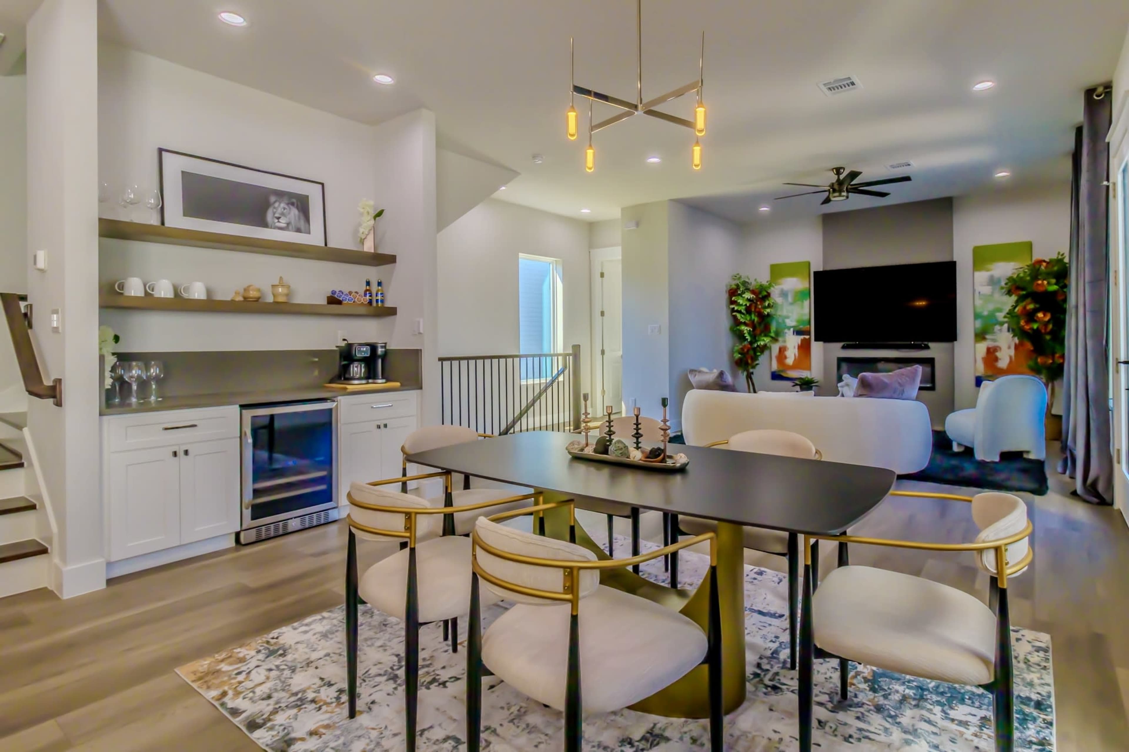 The image shows a modern open-concept dining area featuring a black dining table with light-colored upholstered chairs, a decorative rug, and a living room in the background with a large television and green accents on the walls.