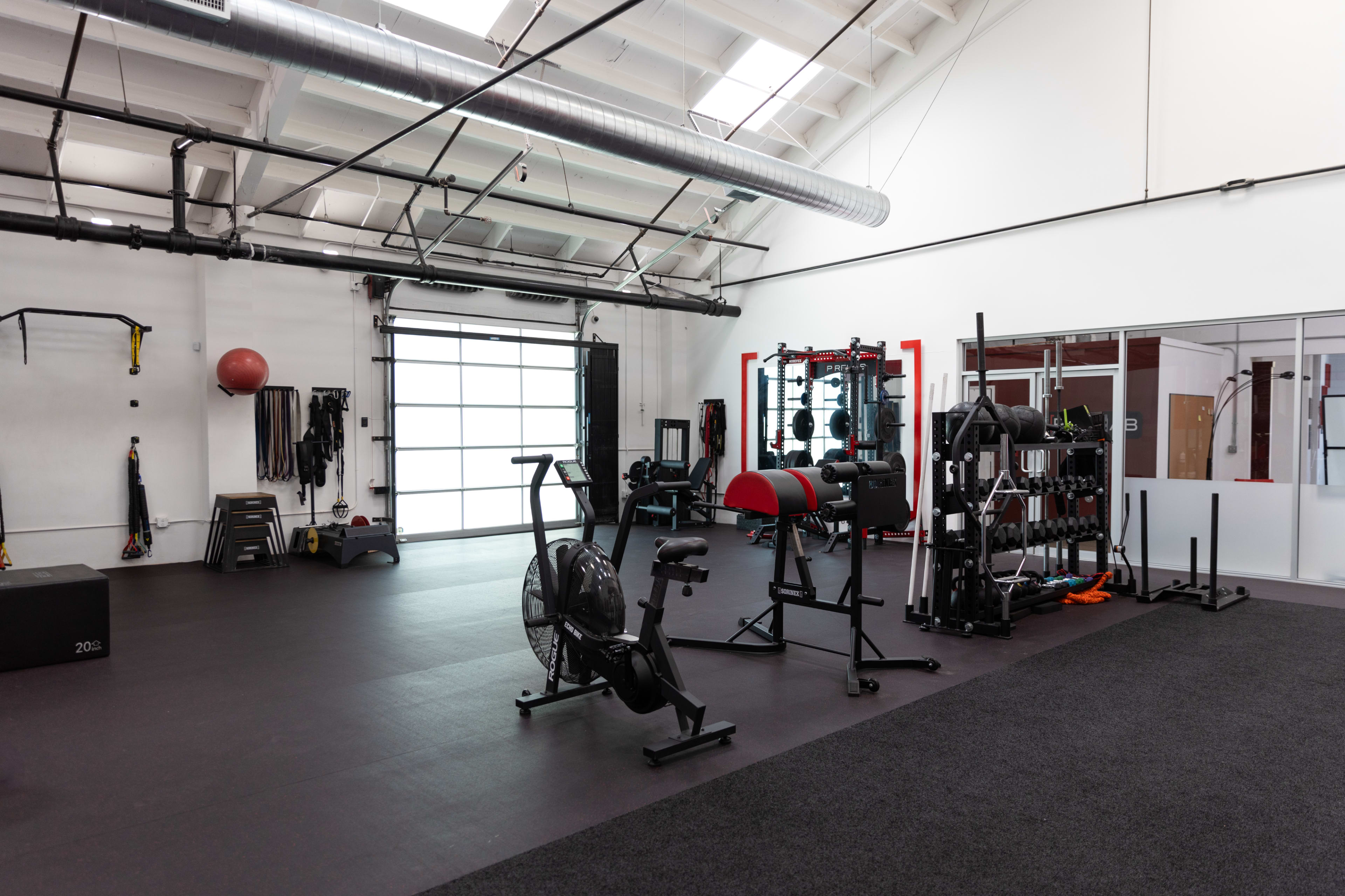 The image shows a spacious gym equipped with various exercise machines and free weights under a high ceiling.