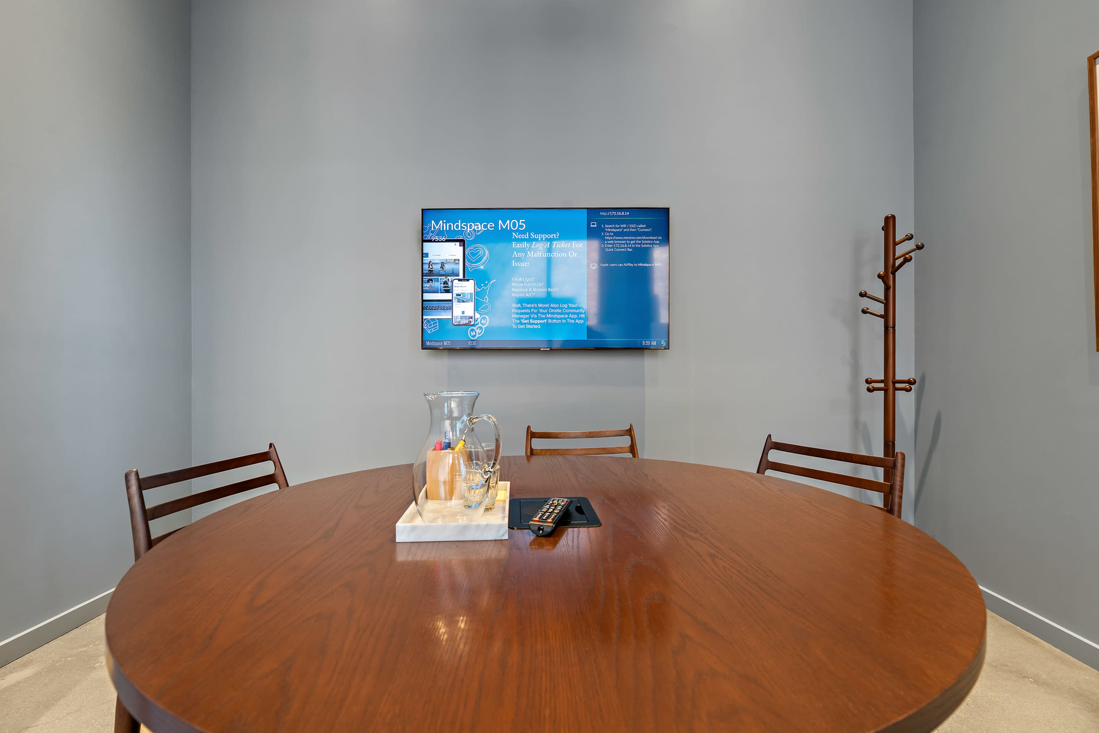 A modern conference room features a round wooden table with chairs, a television mounted on the wall displaying information, and a nearby coat rack.