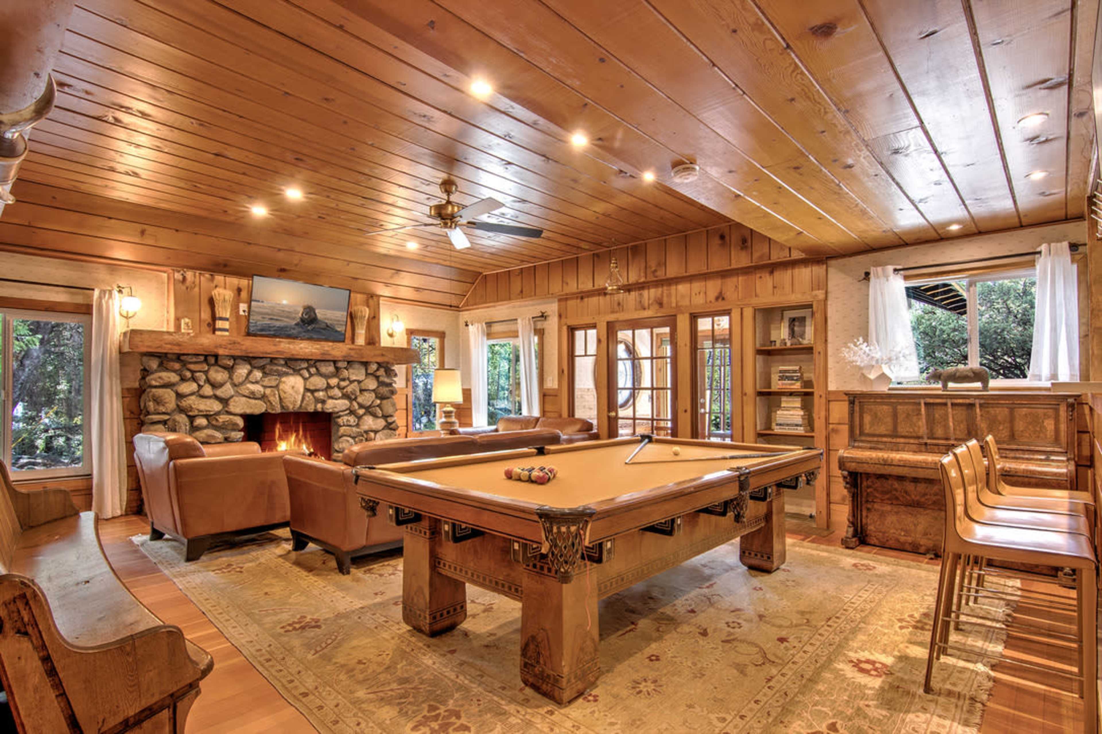 A rustic living room with wood-paneled walls, a stone fireplace, a pool table in the center, and large windows overlooking a green space.