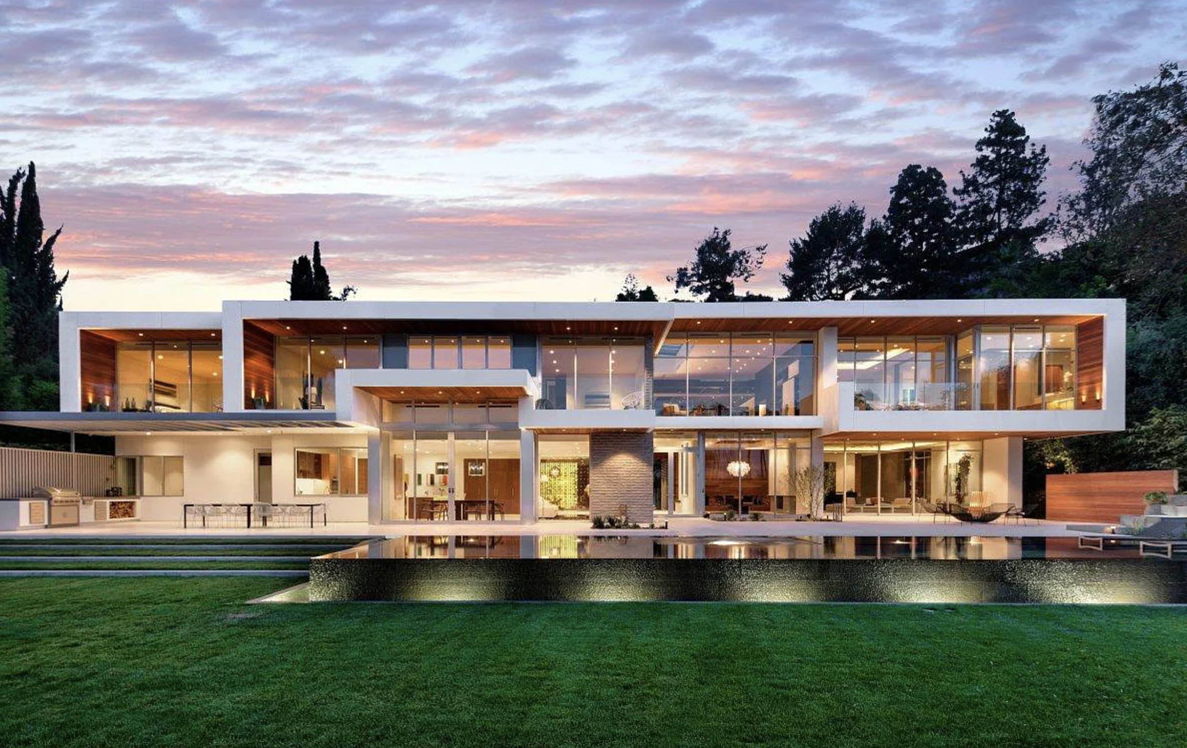 A modern two-story house with large glass windows, a flat roof, and a swimming pool in the foreground reflects the evening sky.