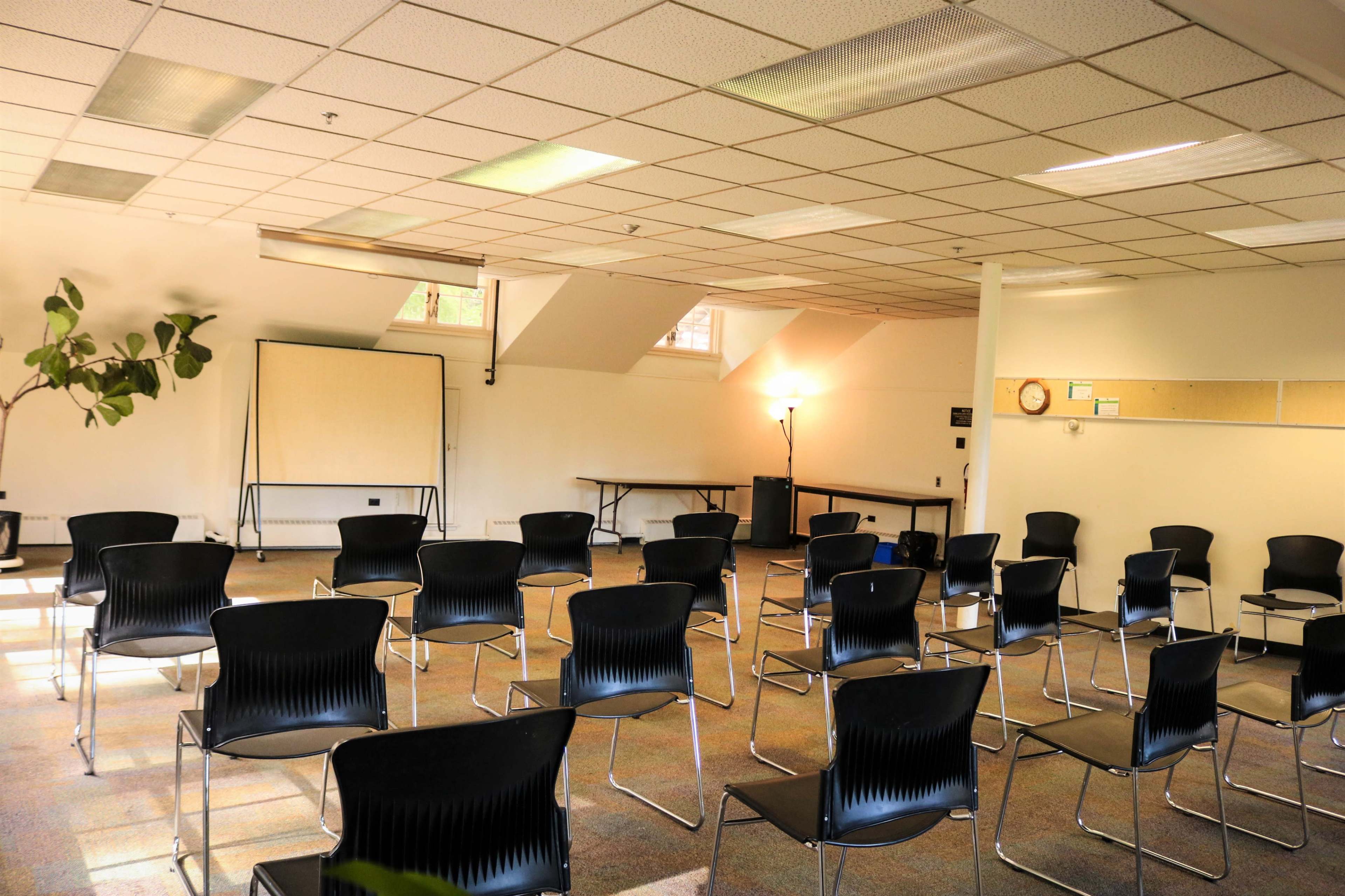 An empty meeting room with rows of black chairs arranged around a small table, a whiteboard on one side, and a large window allowing natural light.
