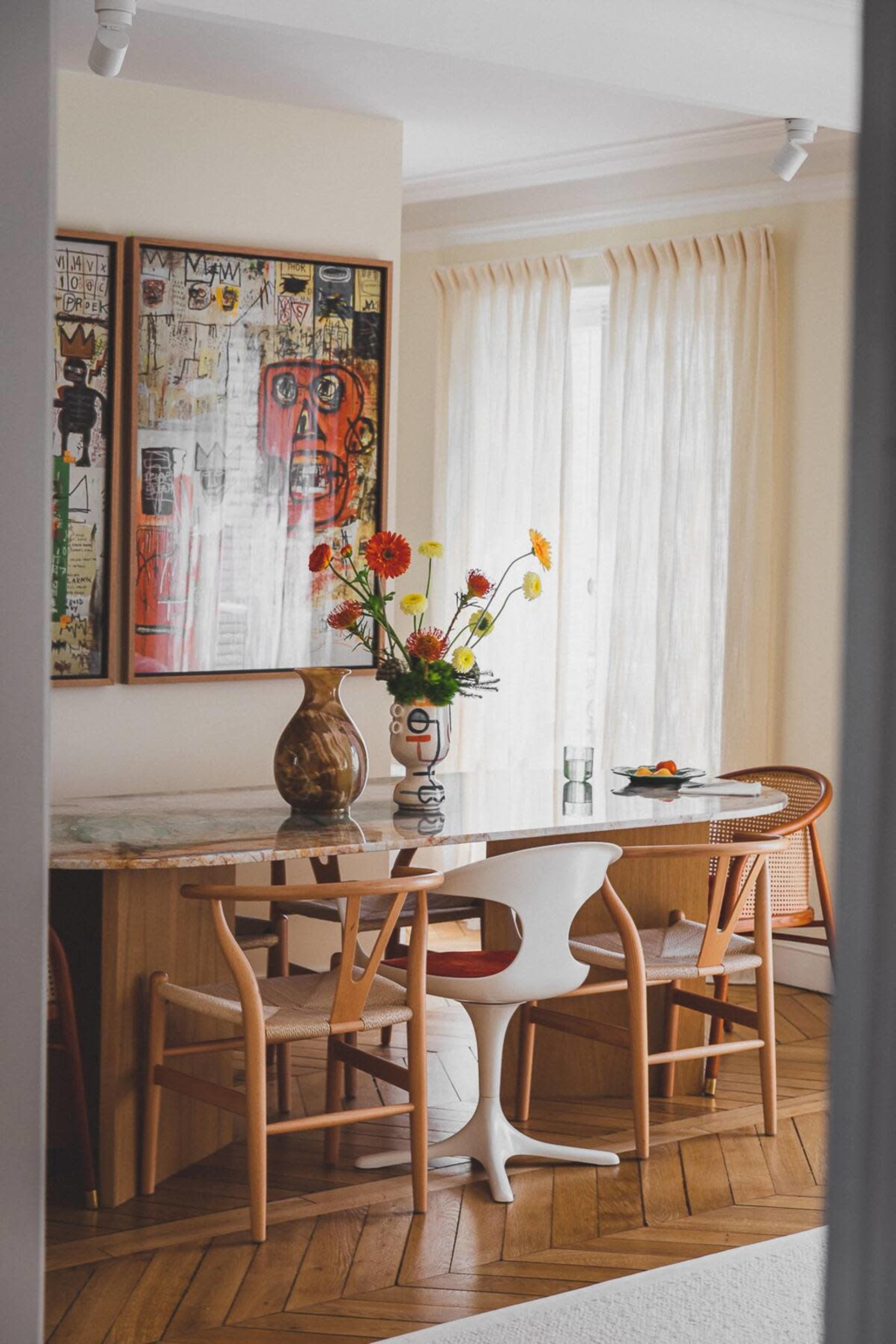 A dining area features a wooden table with a marble top, surrounded by chairs, a vase of flowers, and artwork on the wall.
