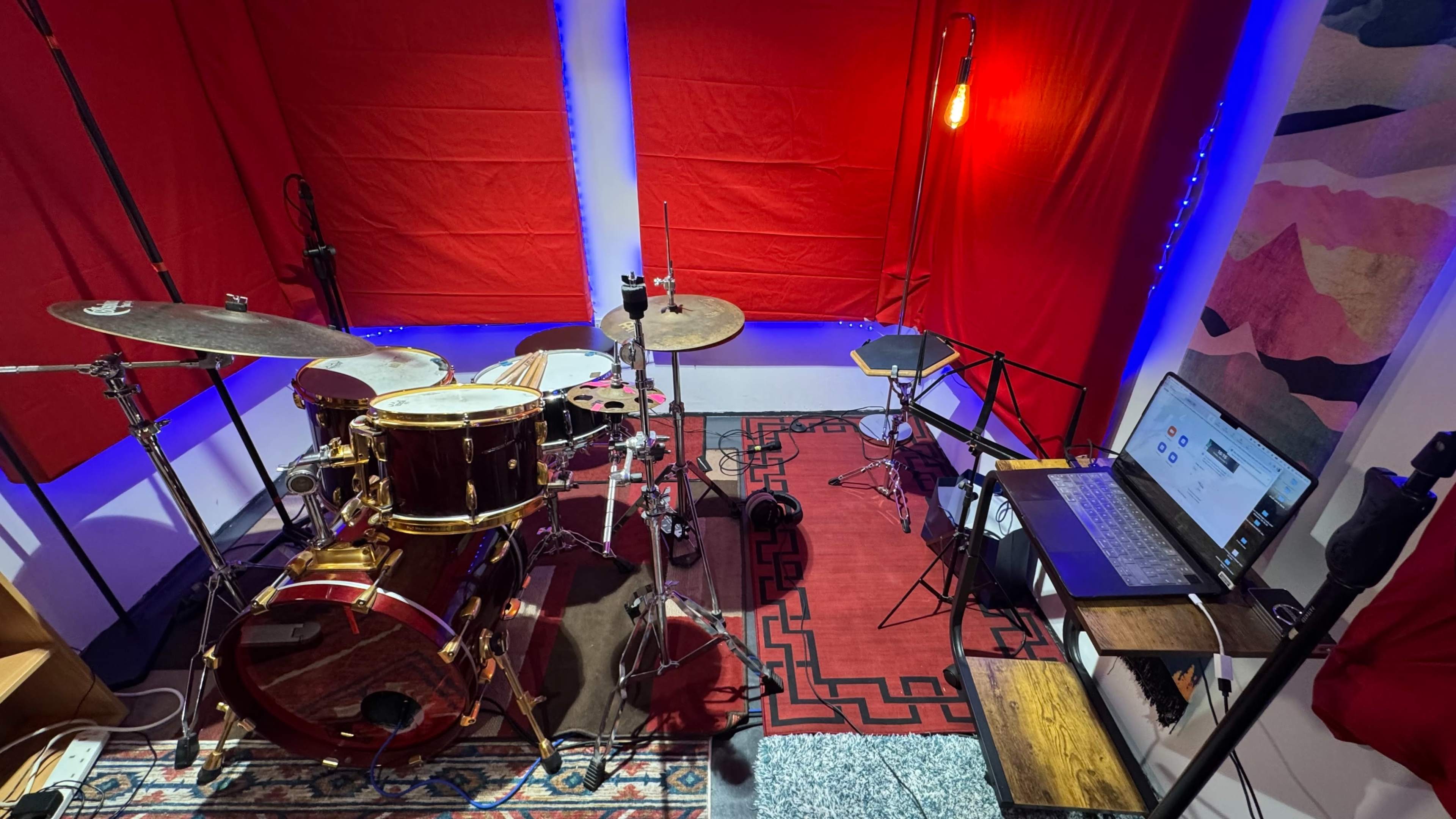 The image shows a music studio setup with a drum kit, a laptop on a wooden table, and red backdrop curtains.