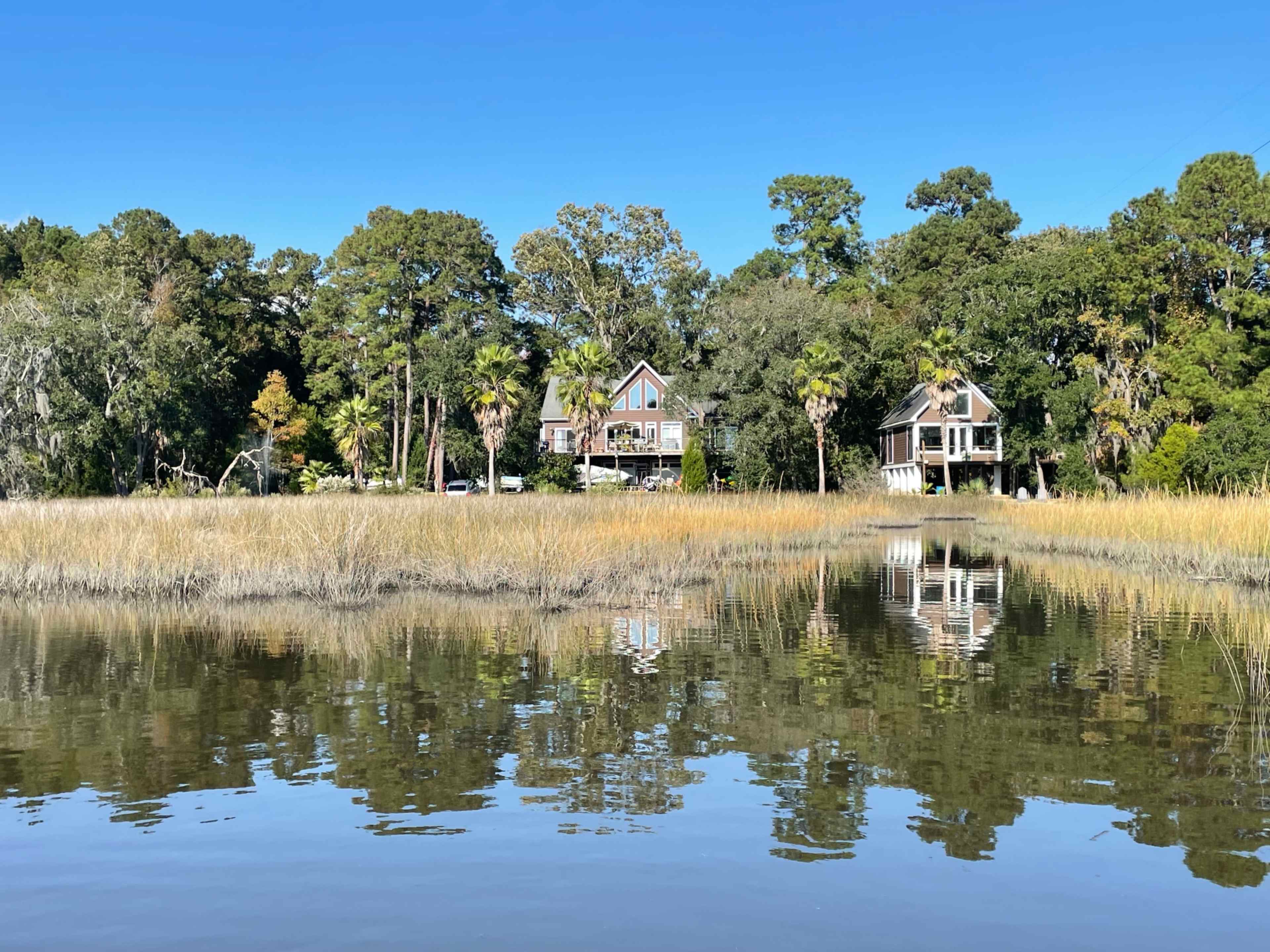 The image shows a calm waterway bordered by tall grasses, with two houses nestled among trees in the background reflecting in the water.