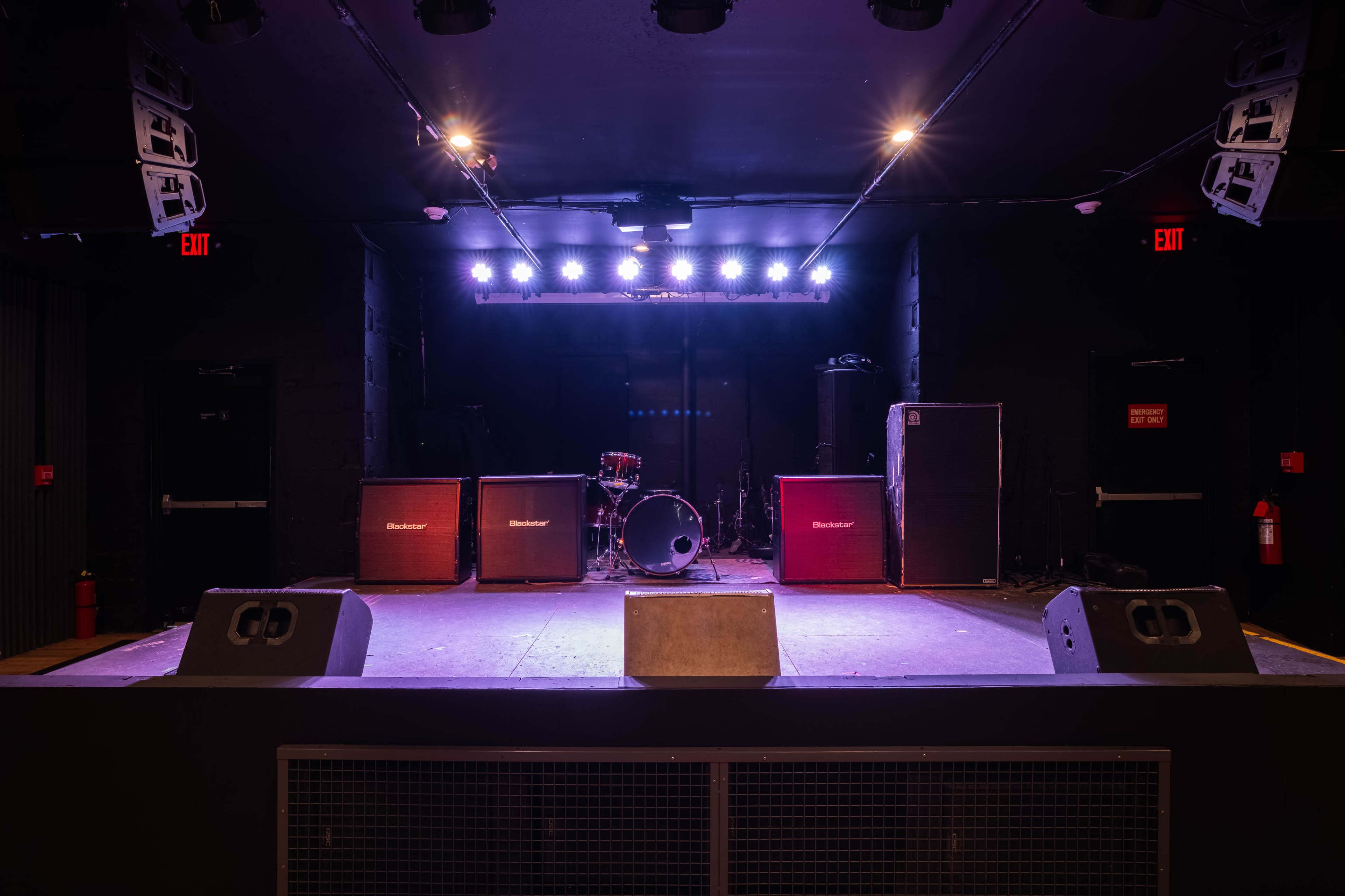 The image shows an empty concert stage with amplifiers, a drum set, and overhead lights.