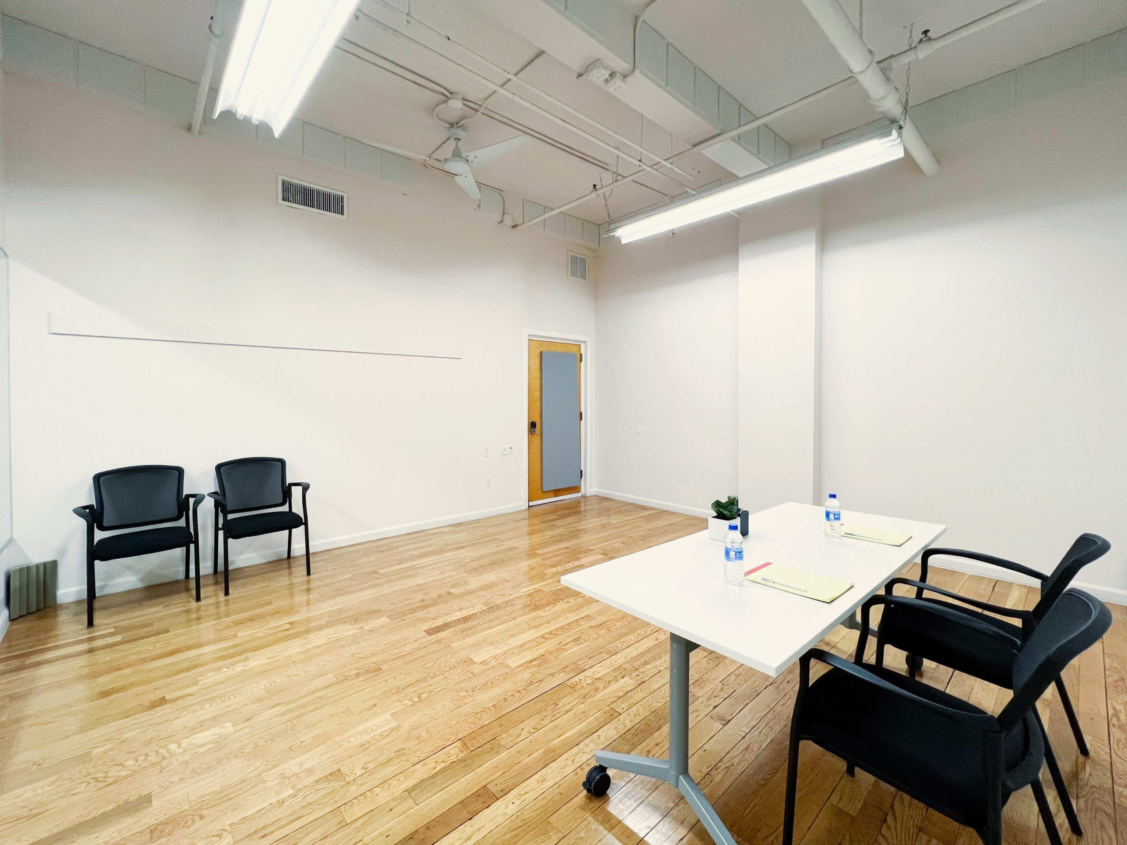 A brightly lit meeting room features a table with two chairs and two water bottles, alongside two additional chairs against the wall.