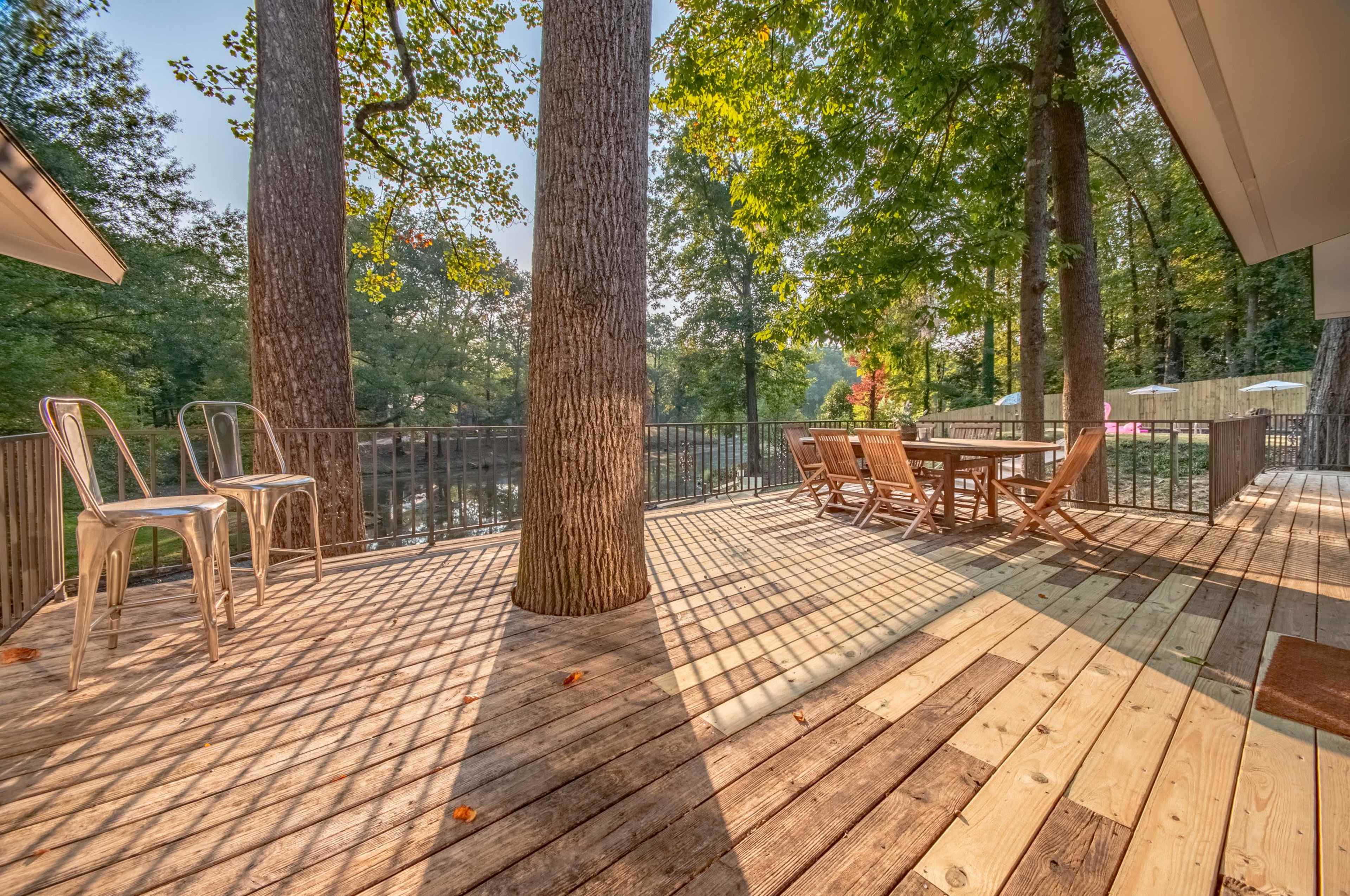 A deck with wooden flooring features metal chairs and a dining table, surrounded by trees overlooking a calm body of water.