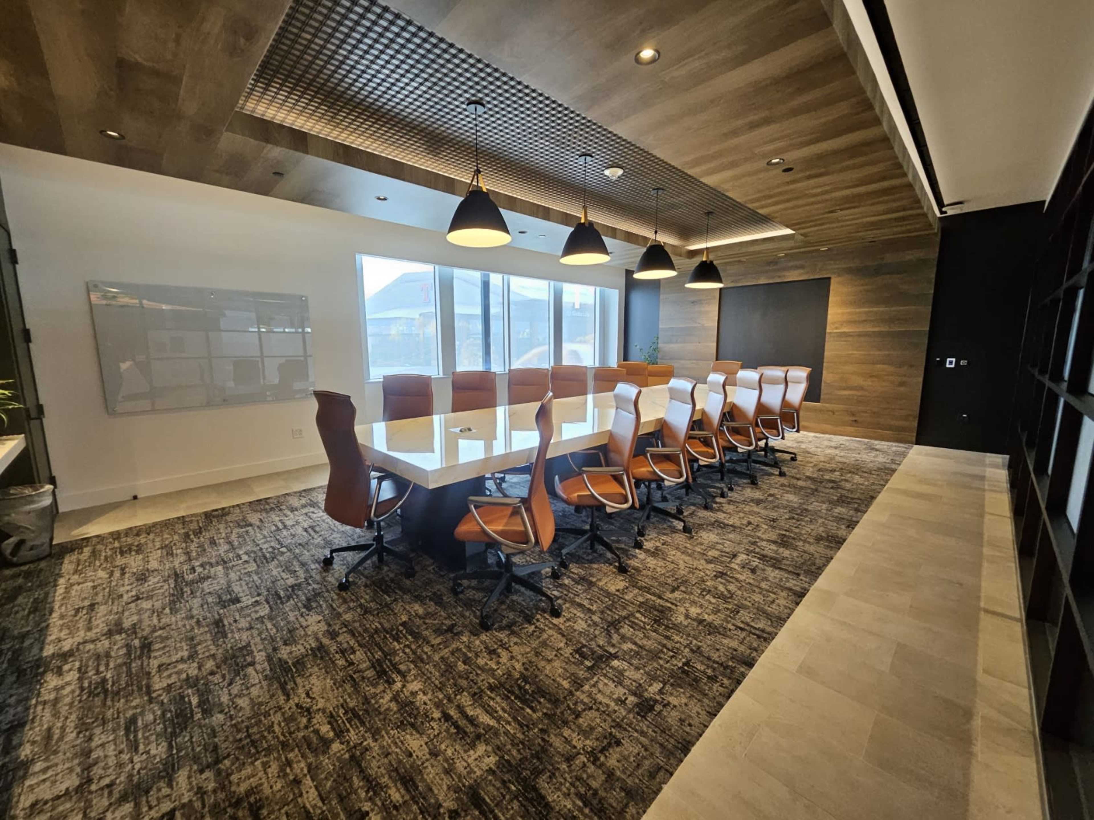 A modern conference room features a long table surrounded by ergonomic orange chairs, with large windows providing natural light and a wooden ceiling overhead.
