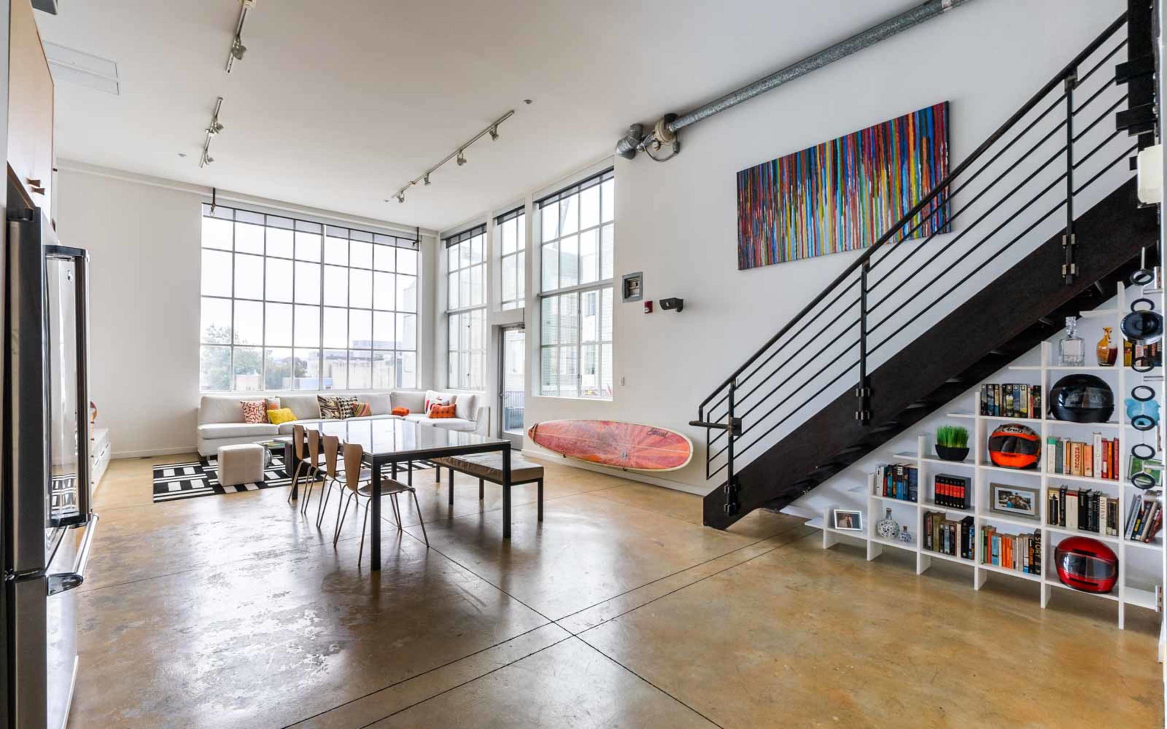 An open-plan living space featuring large windows, a dining area with a table and chairs, a staircase leading to an upper level, and a bookshelf filled with various items.