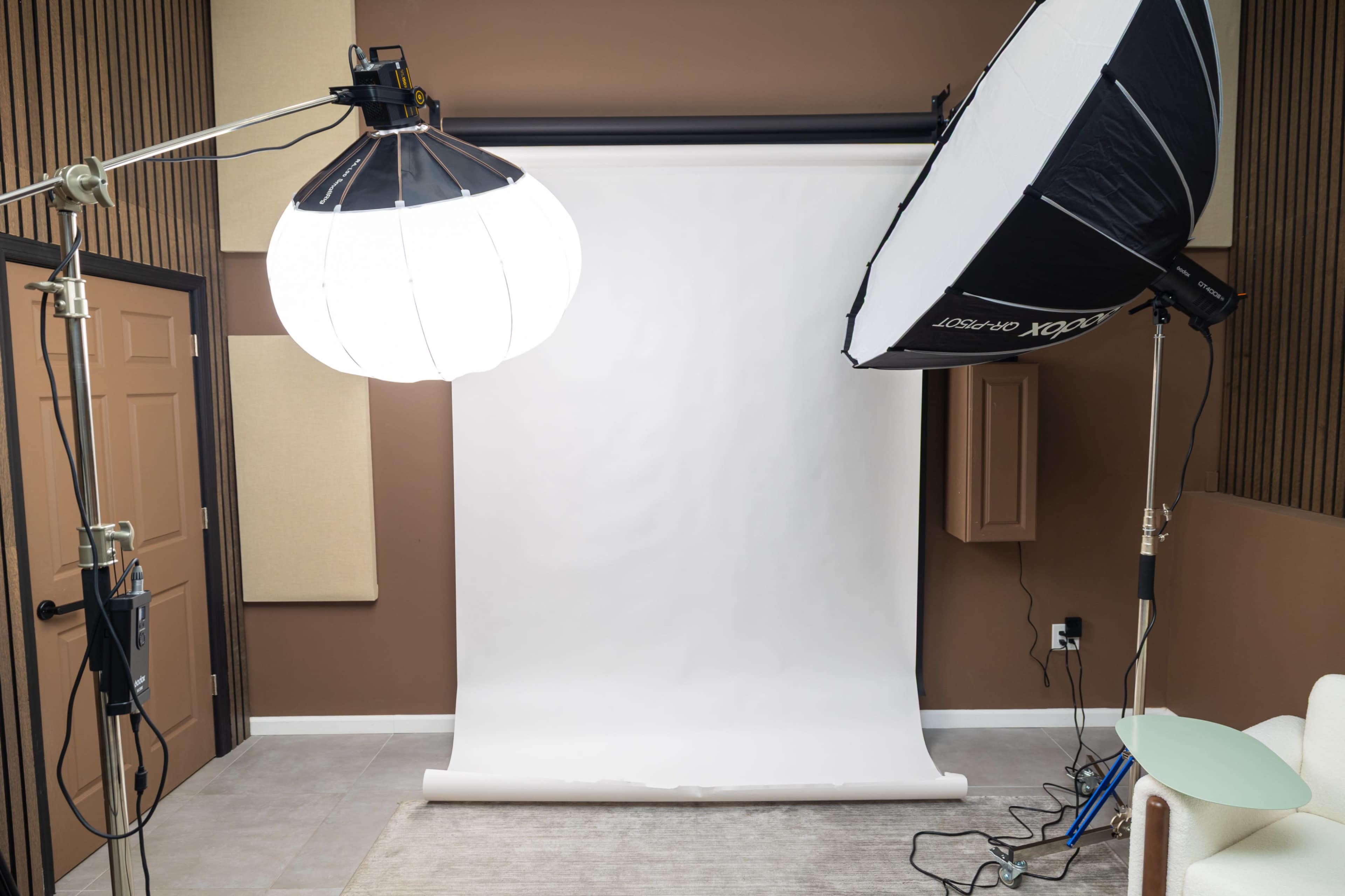 The image shows a photography studio setup with a white backdrop, two lighting equipment, and a neutral-colored floor.