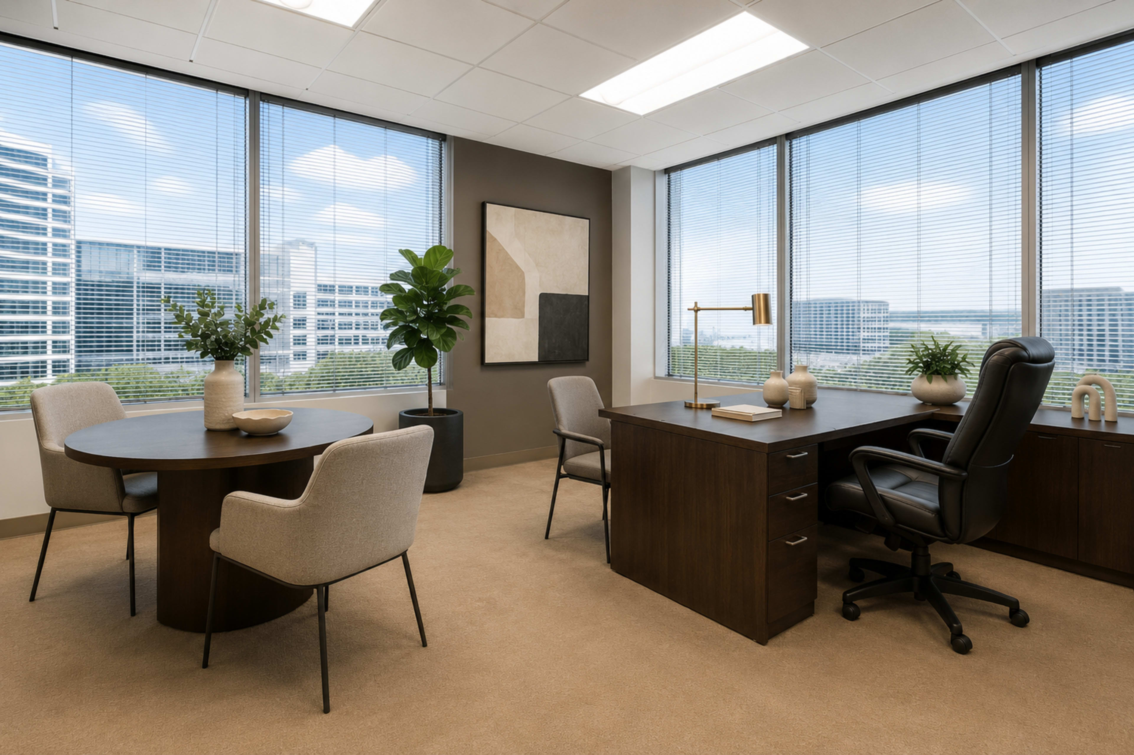 The image shows a modern office space featuring a round table with two chairs, a desk with an office chair, and large windows showcasing a cityscape.