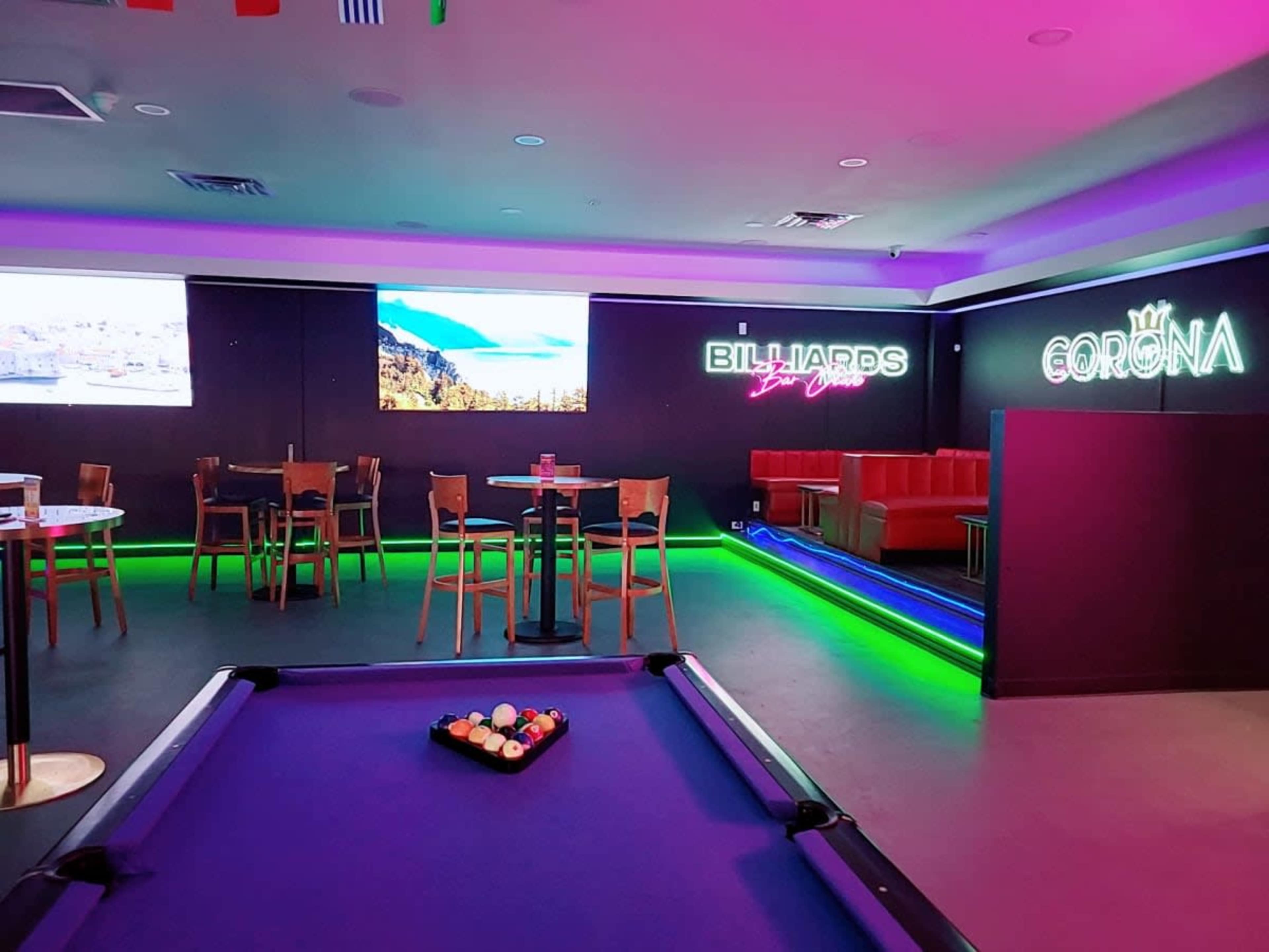 The image shows a modern lounge with a billiards table, colorful lighting, and seating areas featuring tables and chairs.