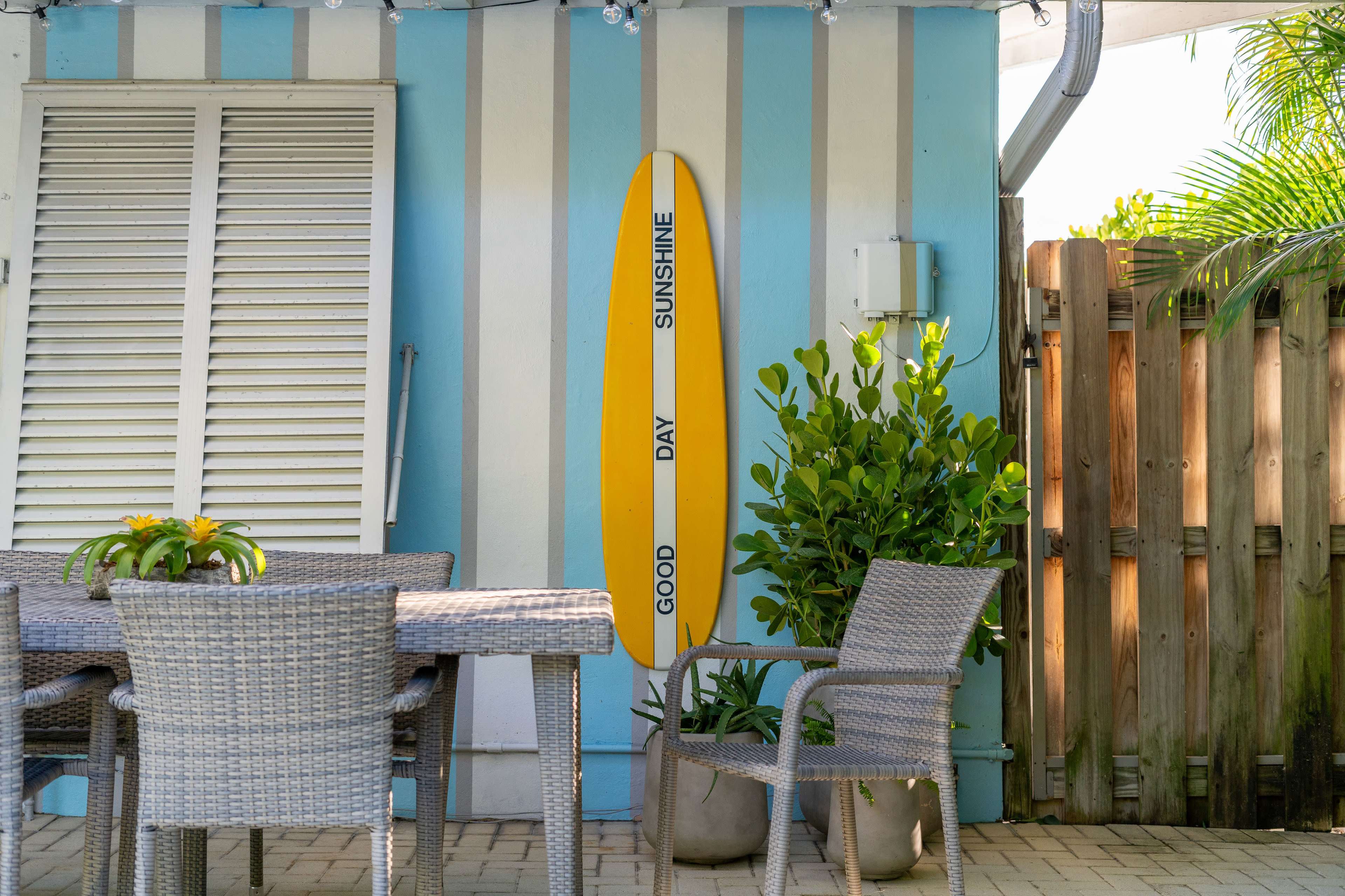 A yellow surfboard with the words "GOOD DAY SUNSHINE" hangs on a blue and white striped wall next to a patio table and chairs surrounded by plants.