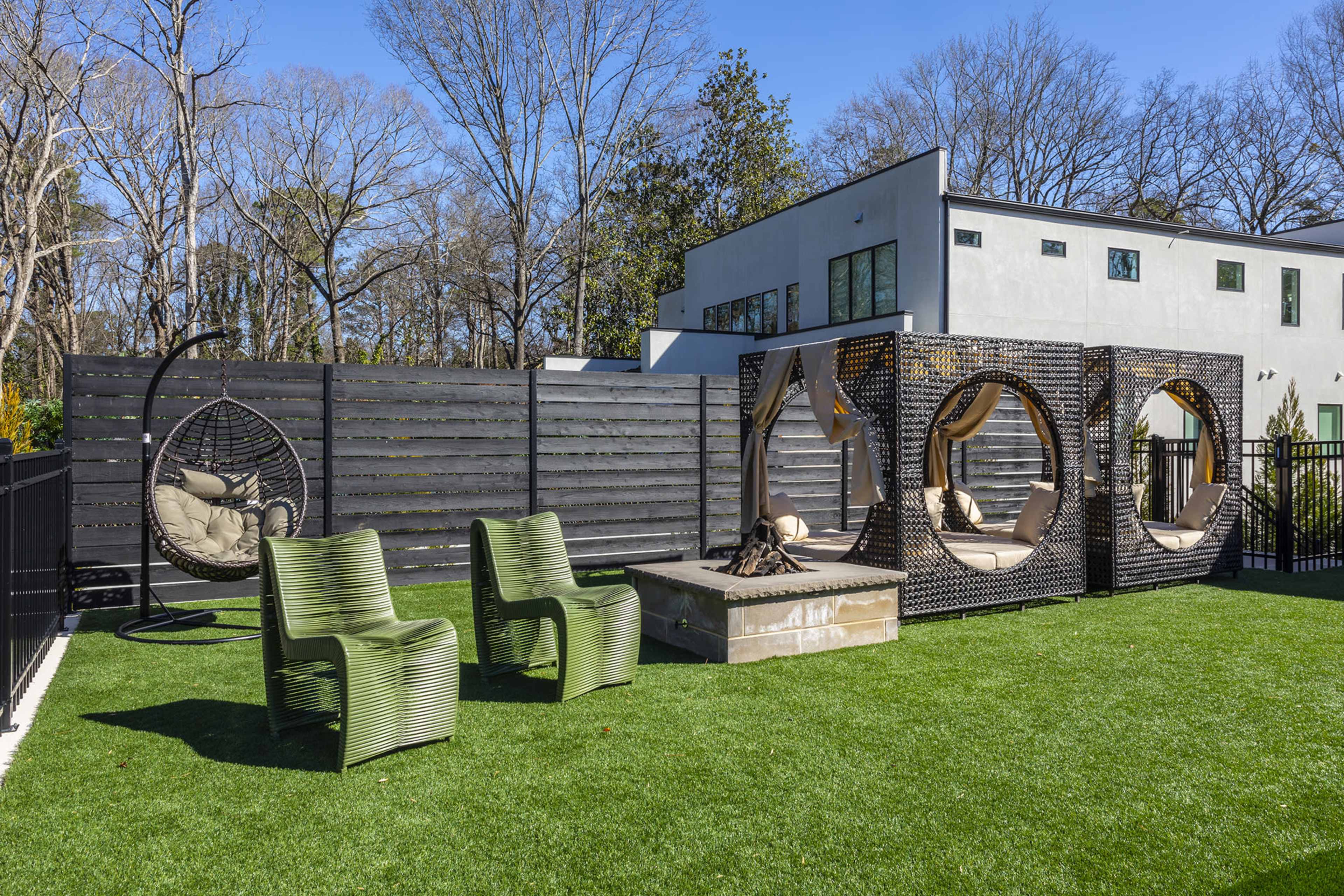 A modern outdoor space featuring two green chairs, a circular hanging chair, and a stone fire pit surrounded by faux grass and a wooden fence.
