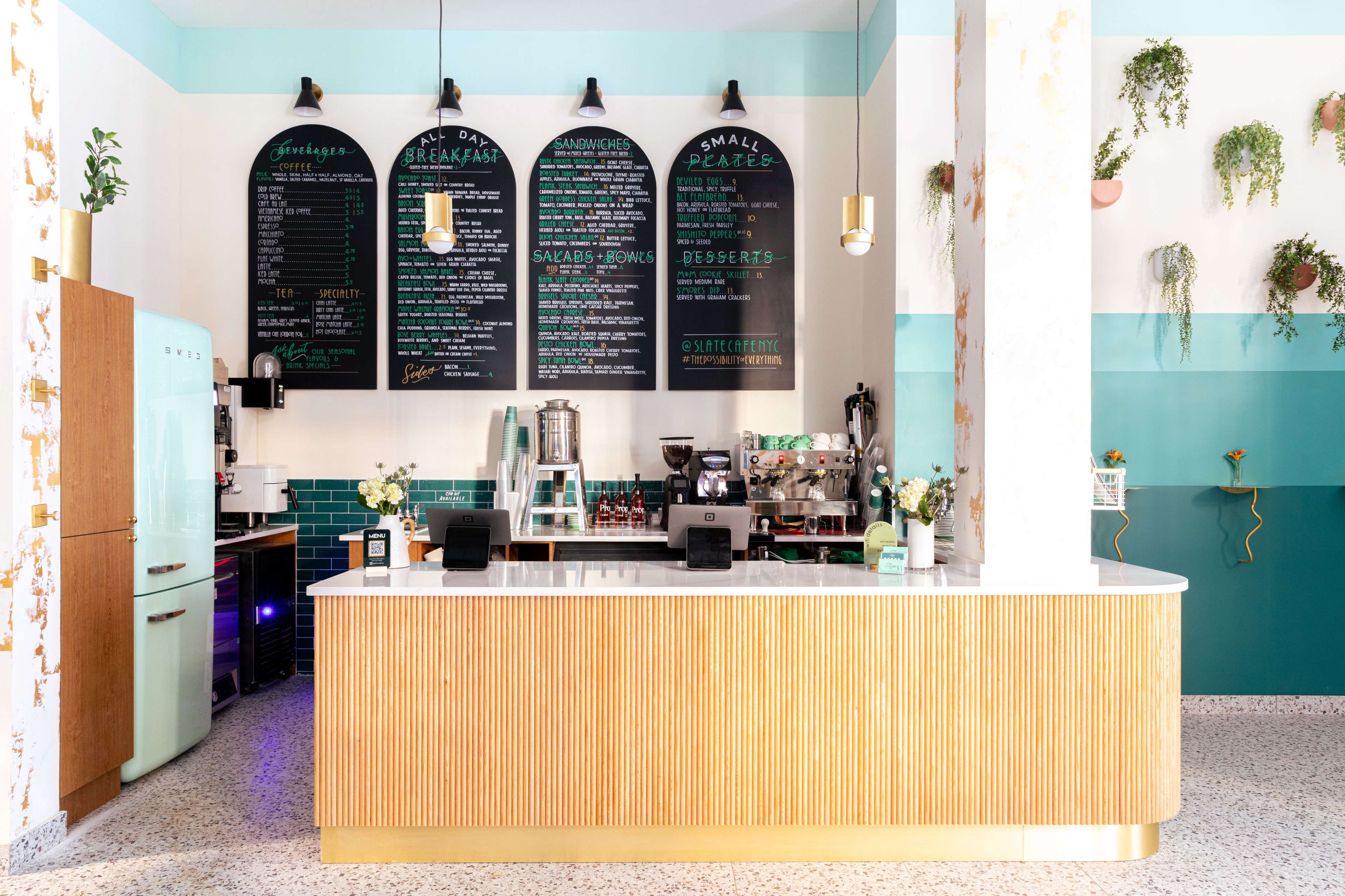 A brightly lit café counter with a menu board above it, featuring a retro-style refrigerator and various plants adorning the walls.