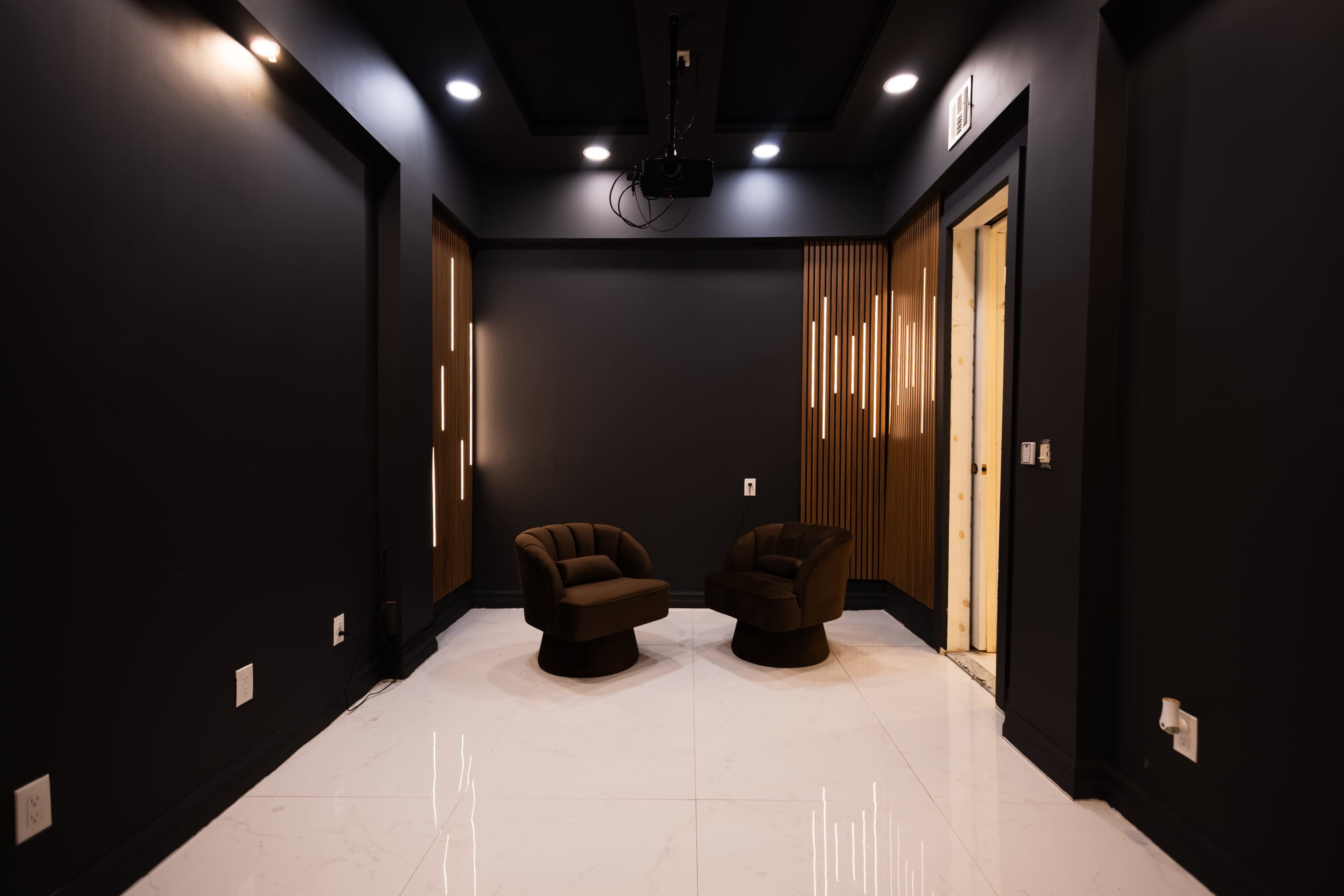 The image shows a modern, minimalist room with two dark armchairs facing each other on a glossy white tiled floor, surrounded by dark walls and accented with wooden paneling.