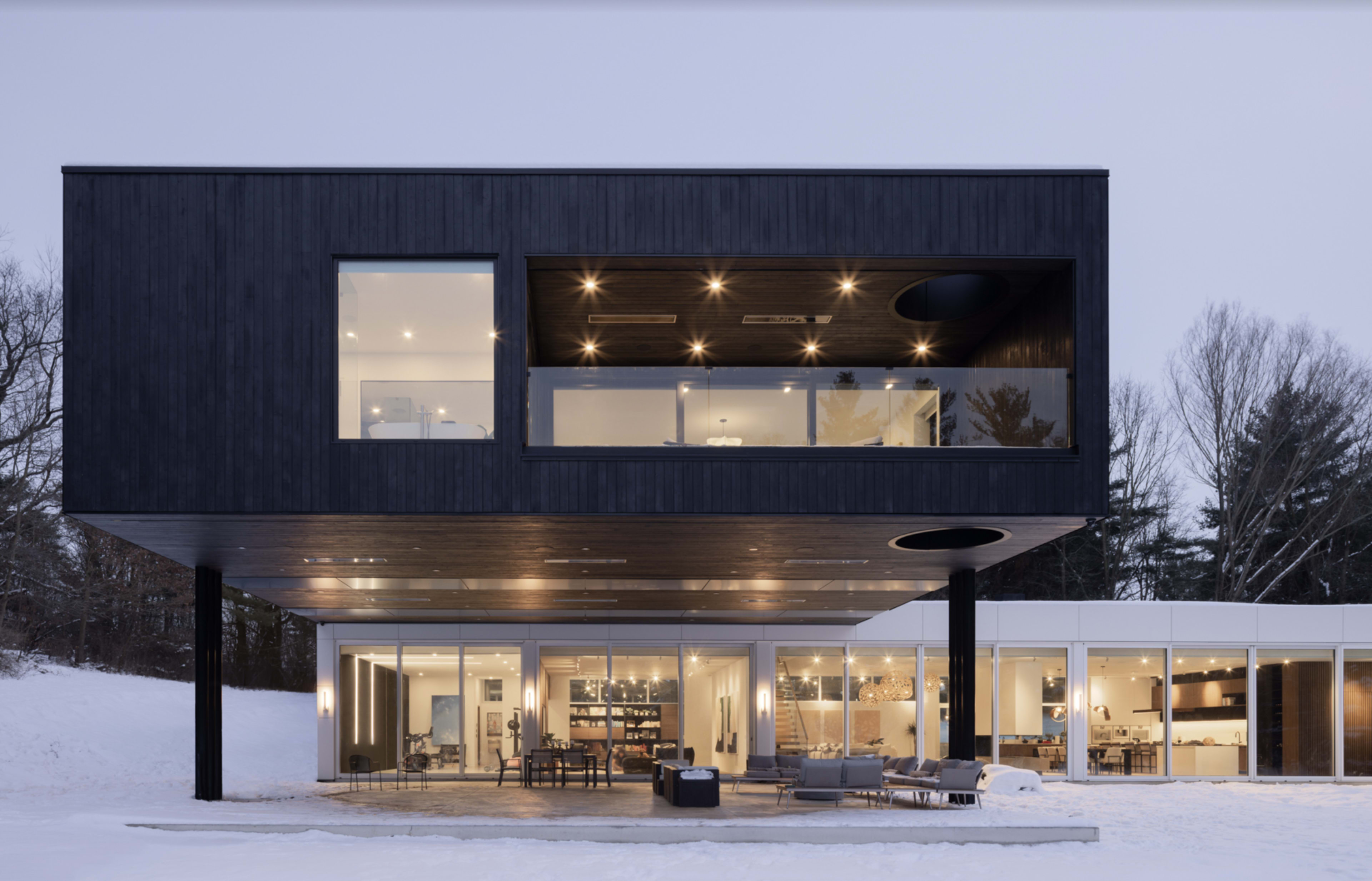 A modern two-story house with a dark wooden exterior and large windows overlooks a snowy landscape.