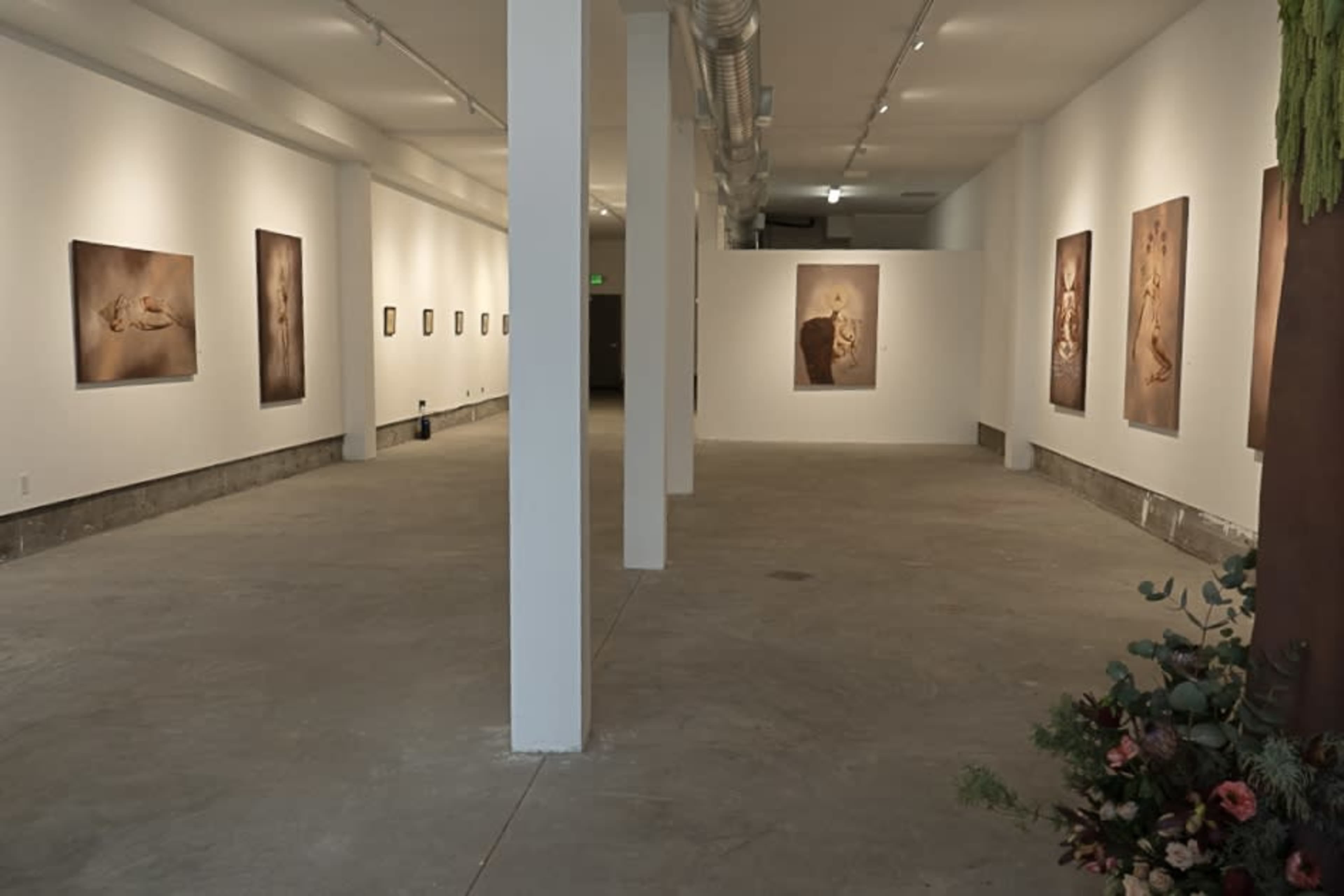 The image shows a spacious art gallery with plain white walls displaying various framed artworks and a concrete floor.