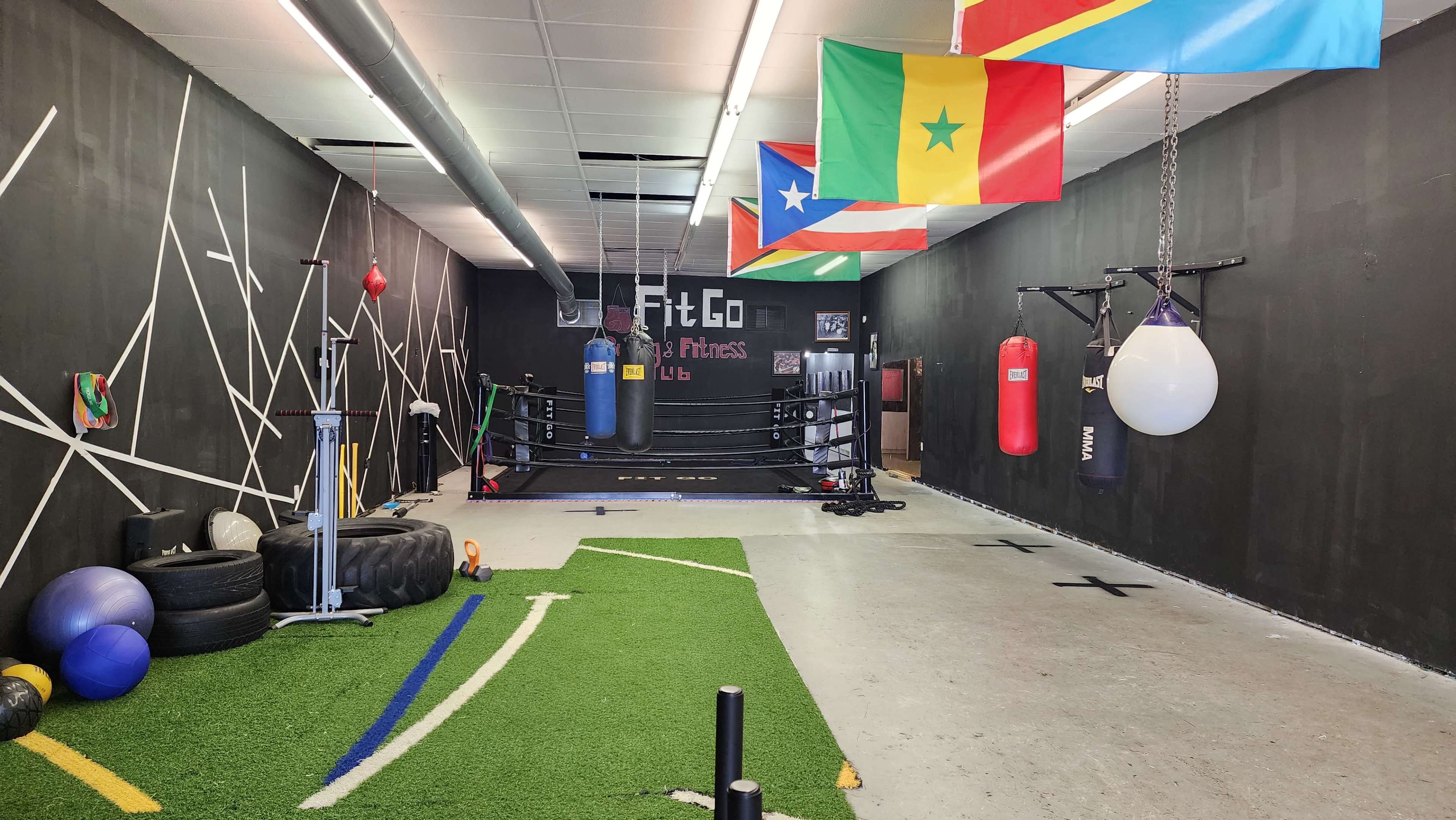 An indoor fitness area with a boxing ring, punching bags, exercise equipment, and various flags hanging on the walls.