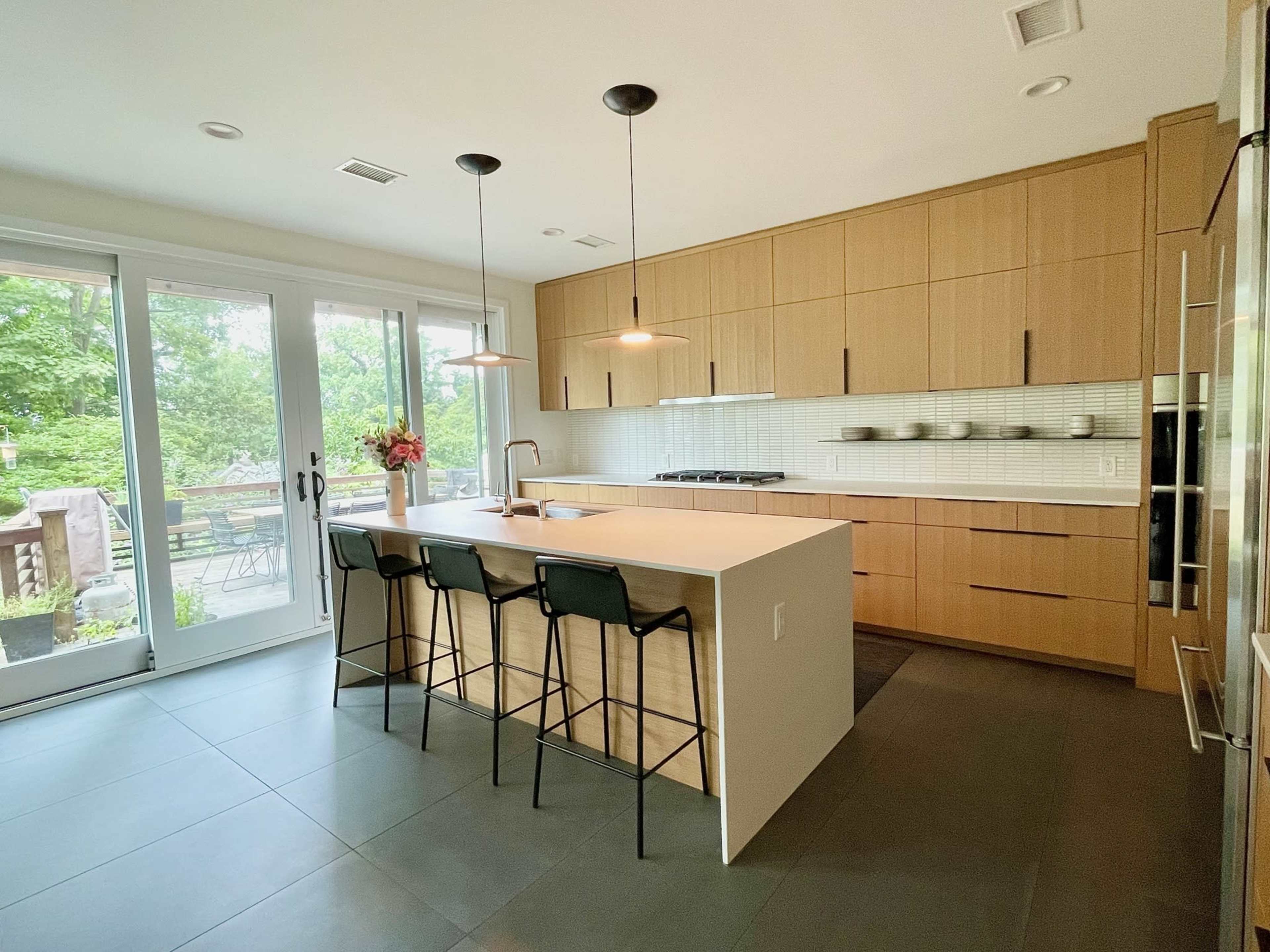 The image shows a modern kitchen with a light wood cabinetry, an island with seating for three, and large windows overlooking a backyard.