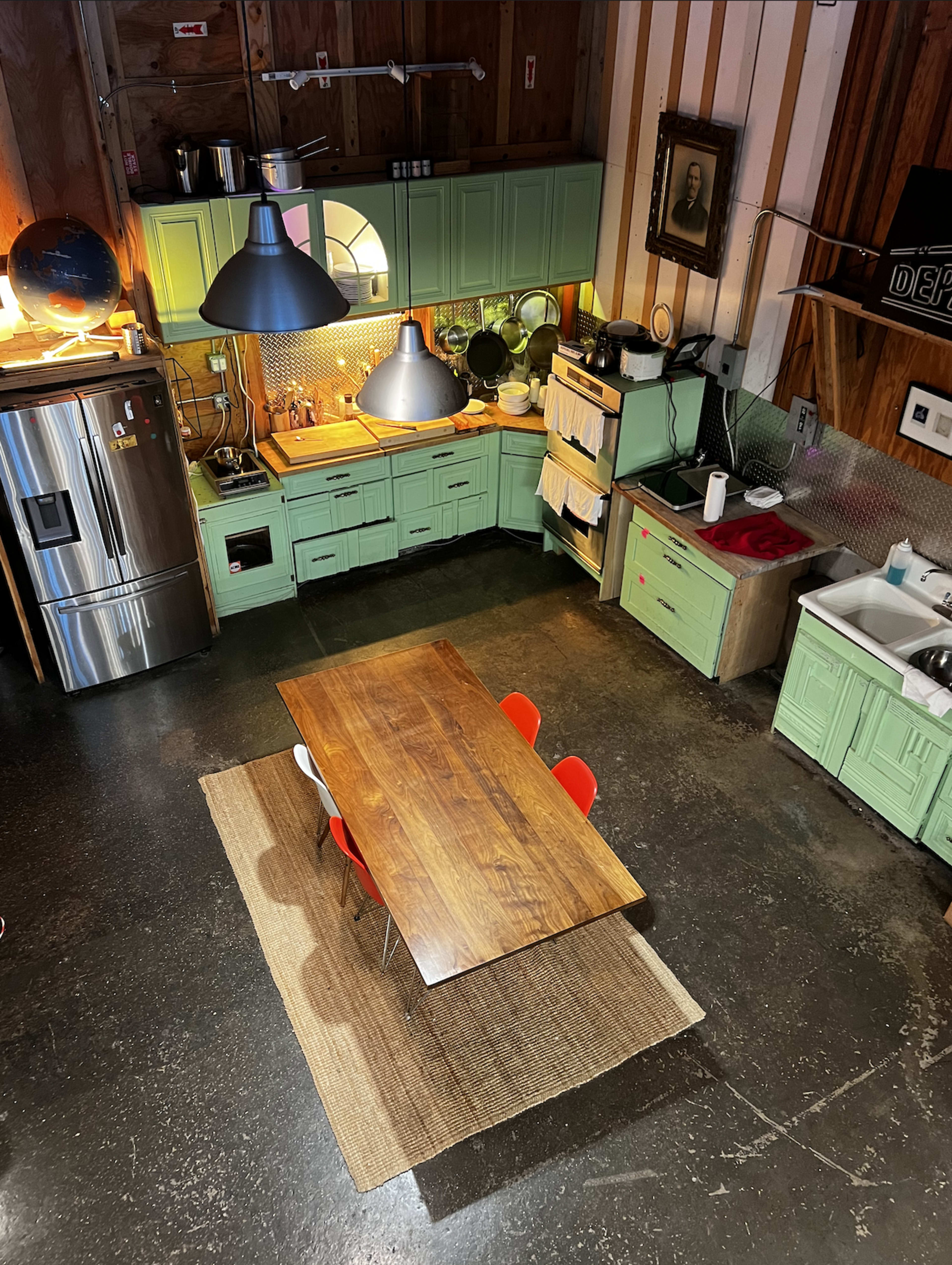 The image shows a modern kitchen with green cabinets, a wooden dining table surrounded by red chairs, and various appliances against a wooden wall.