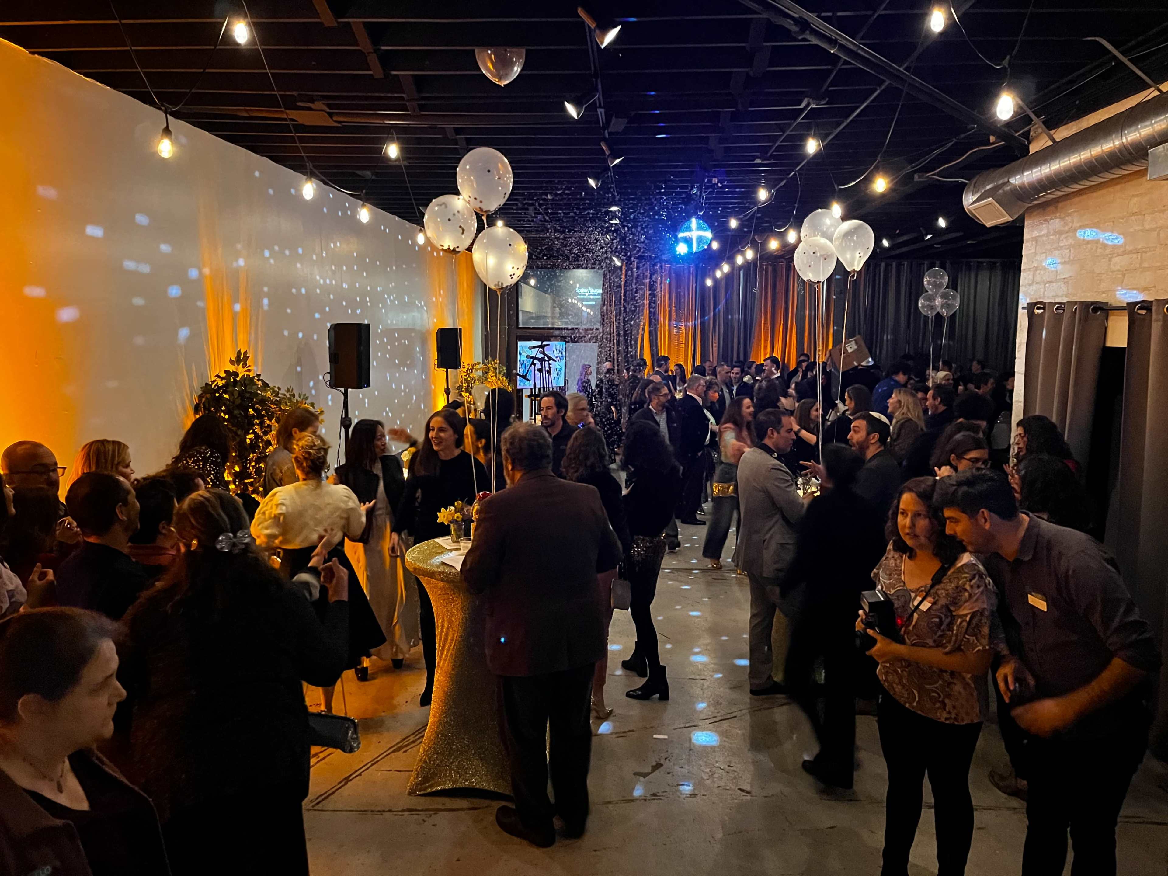 A large crowd of people gathers in a spacious event hall decorated with balloons and string lights, celebrating an occasion.