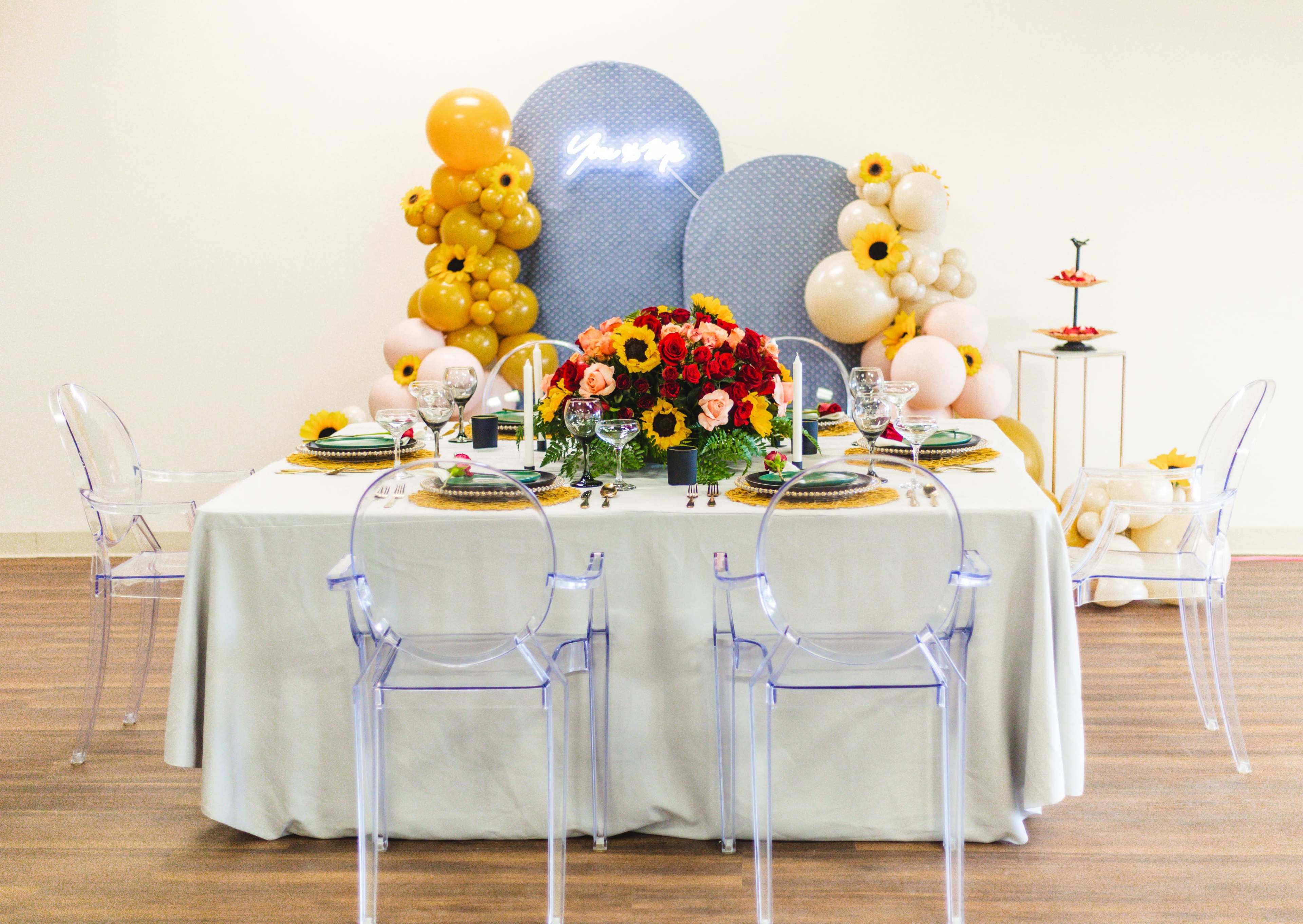 A dining table is elegantly set with flowers, colorful dishes, and decorations, surrounded by clear acrylic chairs against a backdrop of balloons and a neon sign.