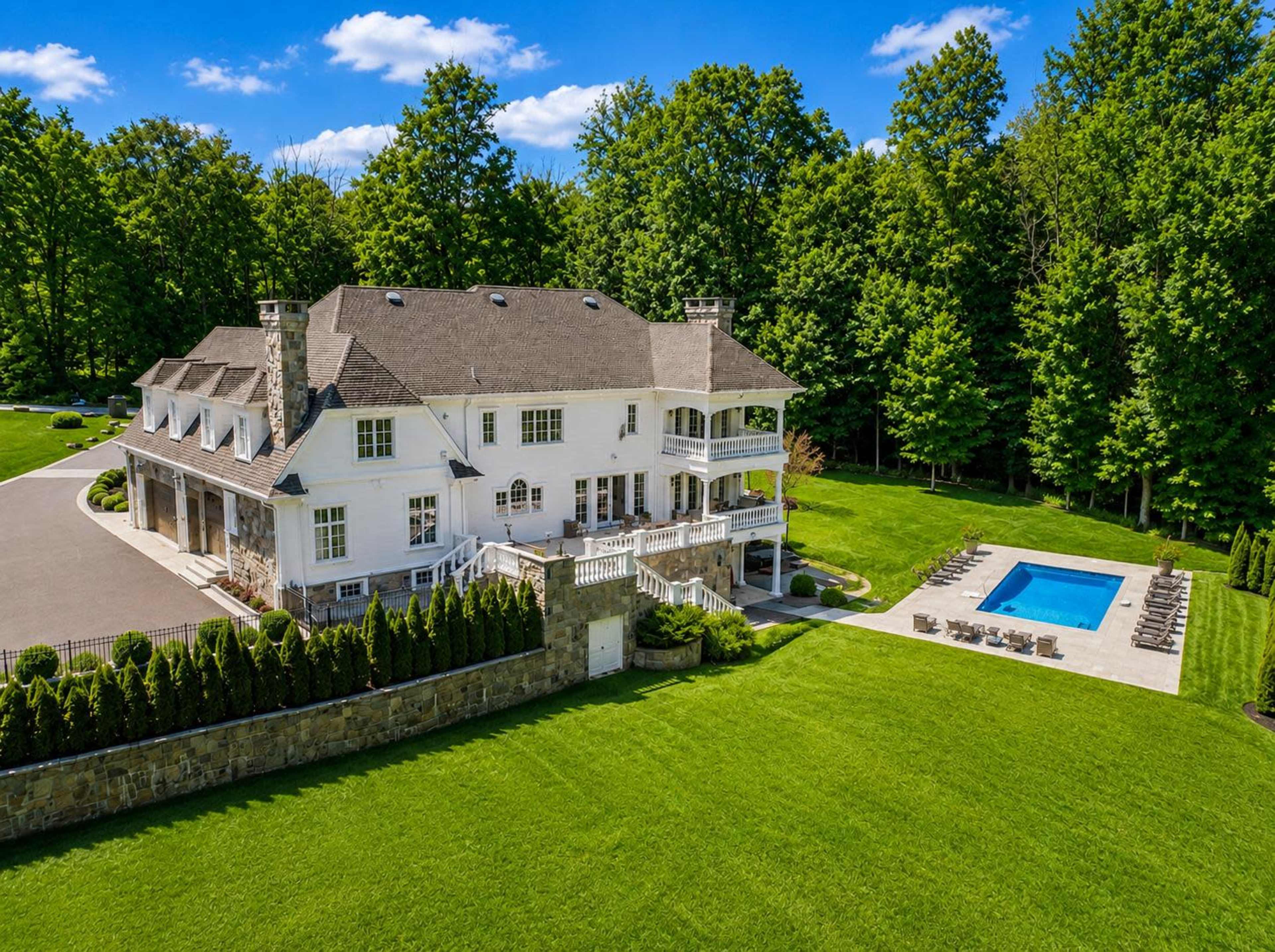 The image shows a large white house with multiple balconies and a pool in the backyard, surrounded by lush green trees.