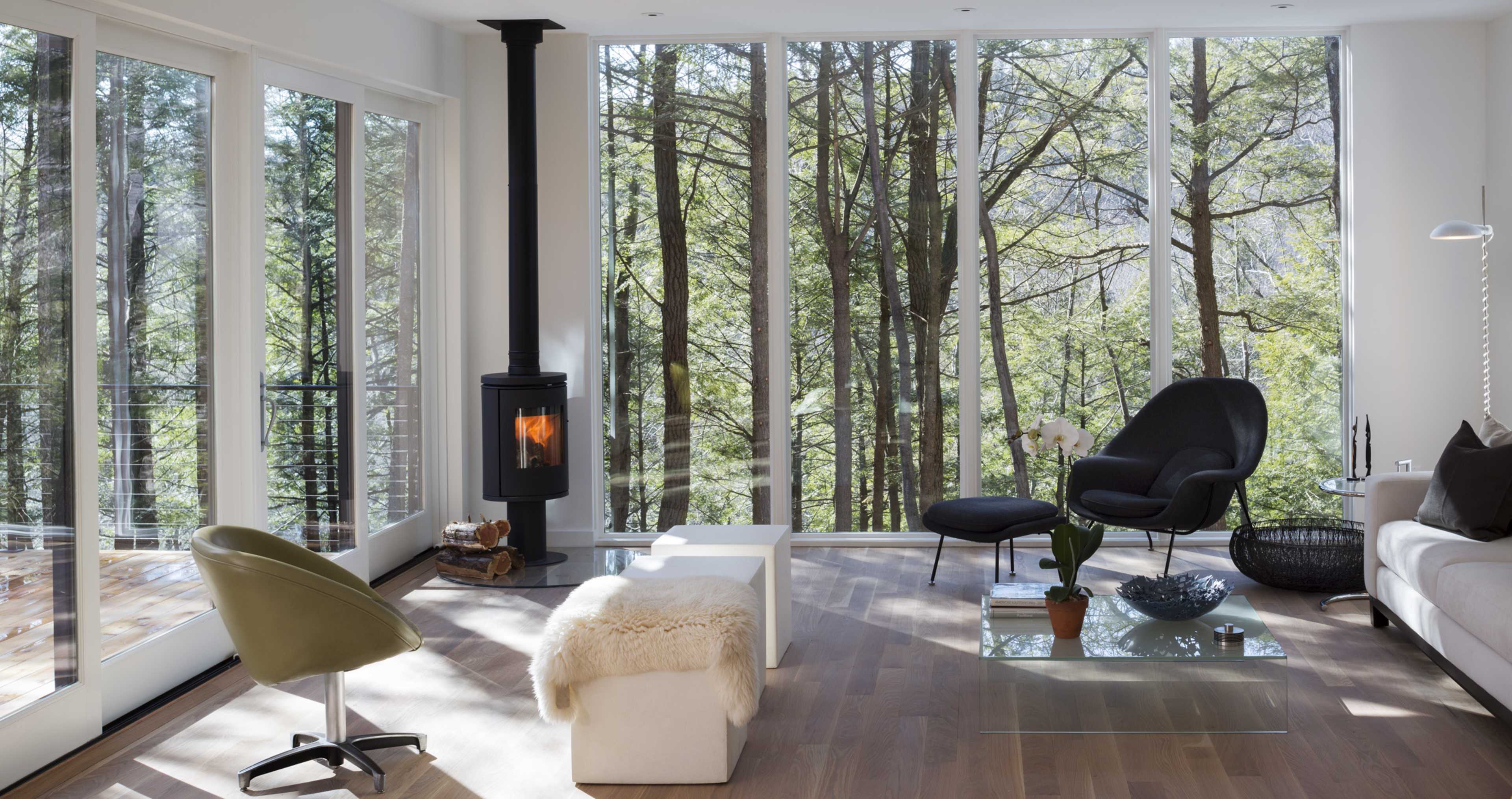 The living room features large windows overlooking a wooded area, with a modern fireplace, stylish furniture, and a minimalist design.